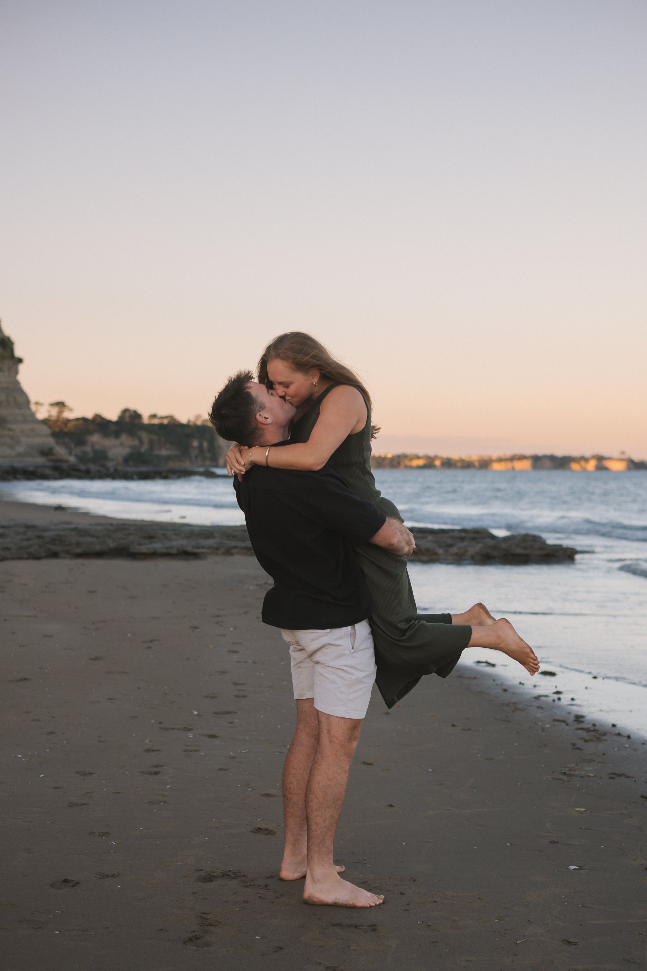 A man holding a woman in a joyful embrace on the beach during sunset, with cliffs and water in the background.