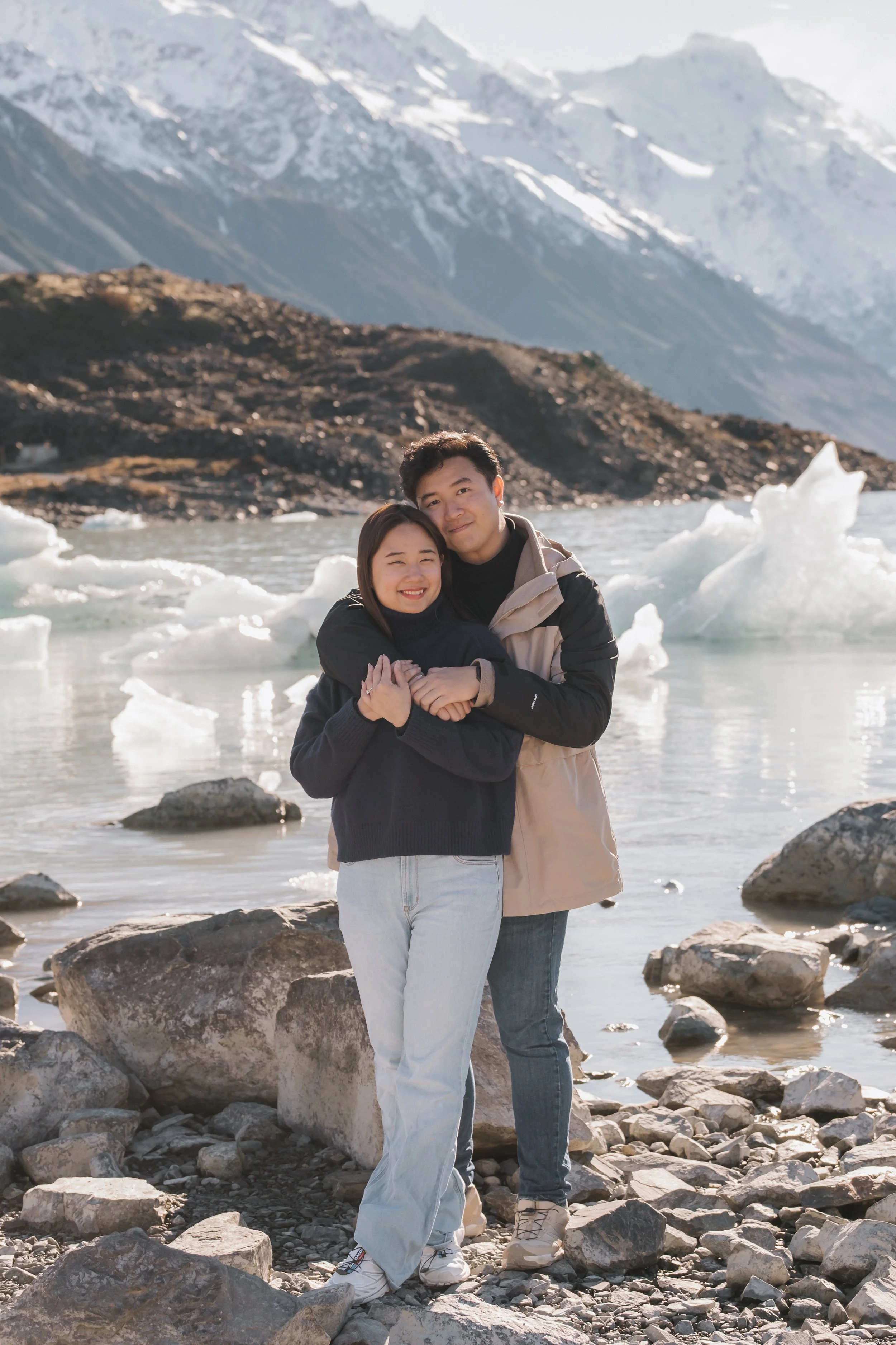 A young couple standing on rocky terrain by a glacier-fed lake with snow-capped mountains in the background. They are hugging and smiling.