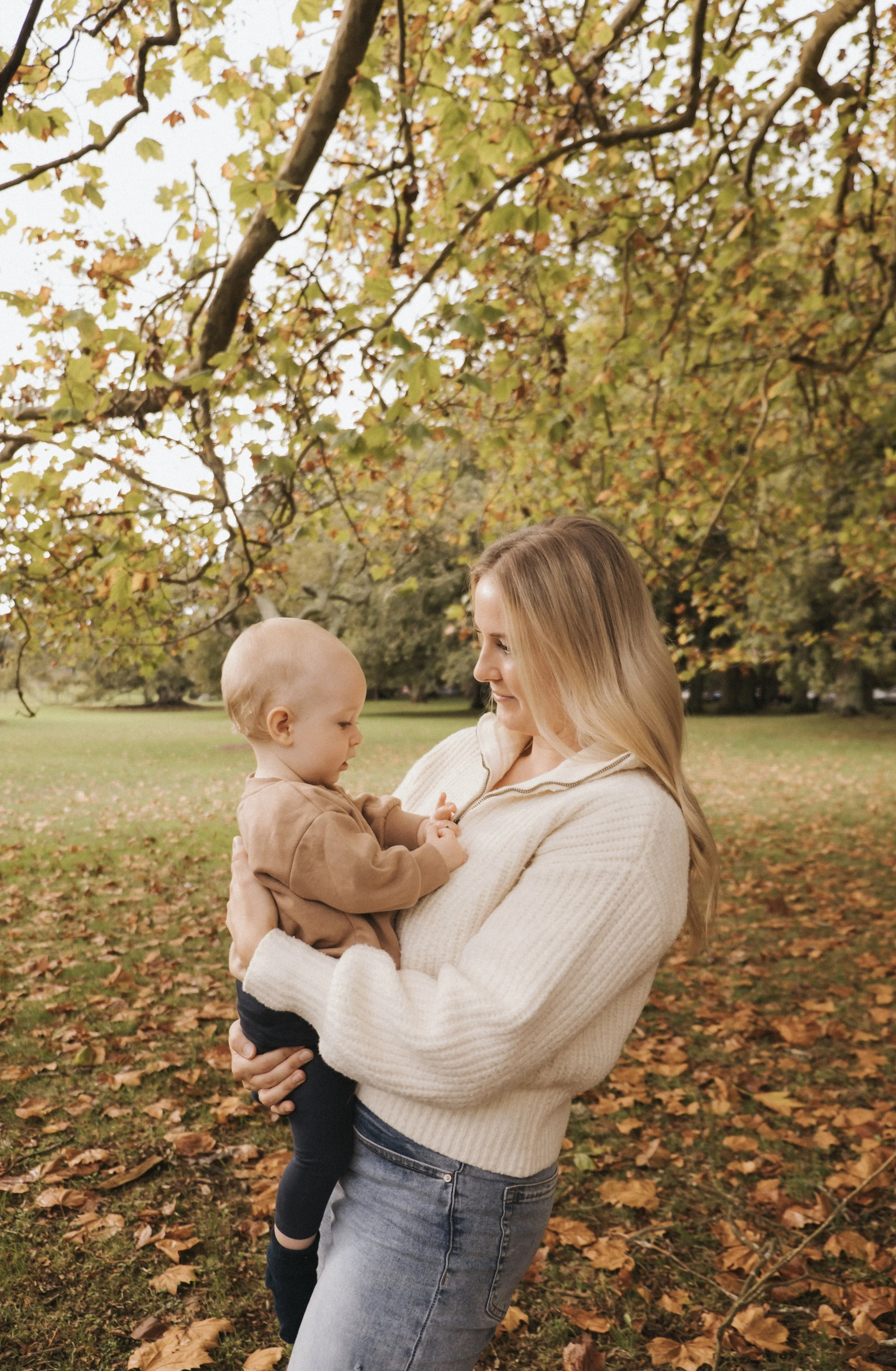 Woman holding a baby in a park with autumn leaves and trees.