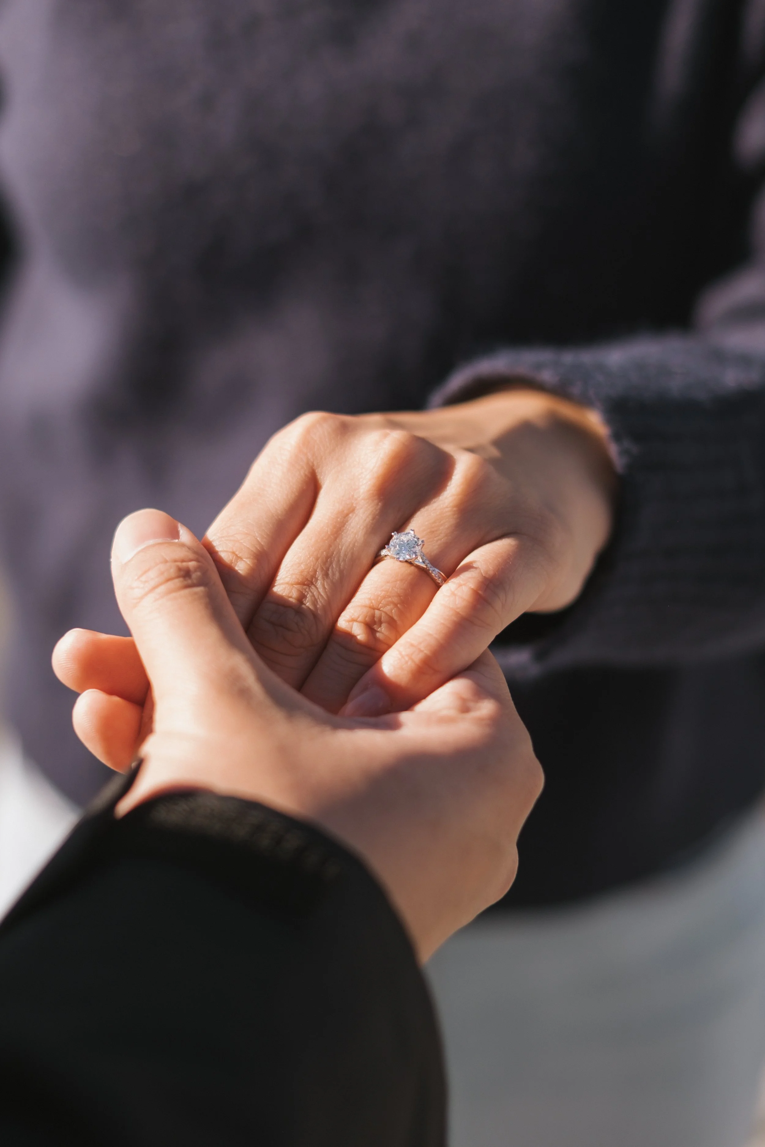 A person is holding another person's hand, which is wearing a diamond engagement ring. The background is blurred, emphasizing the hands and ring.