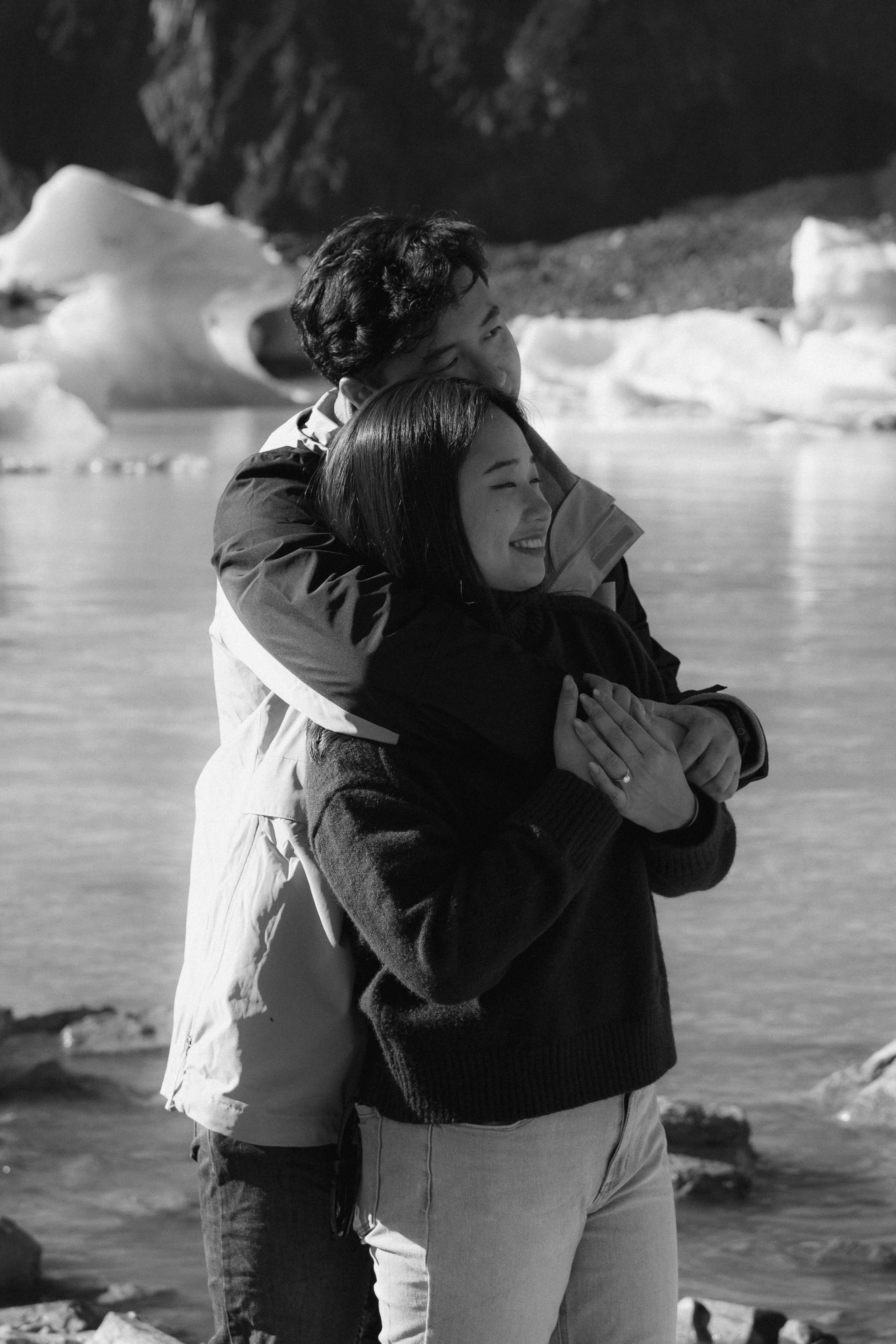 Two people, a man and a woman, embracing each other on a rocky shoreline with icebergs in the background, black and white photograph.