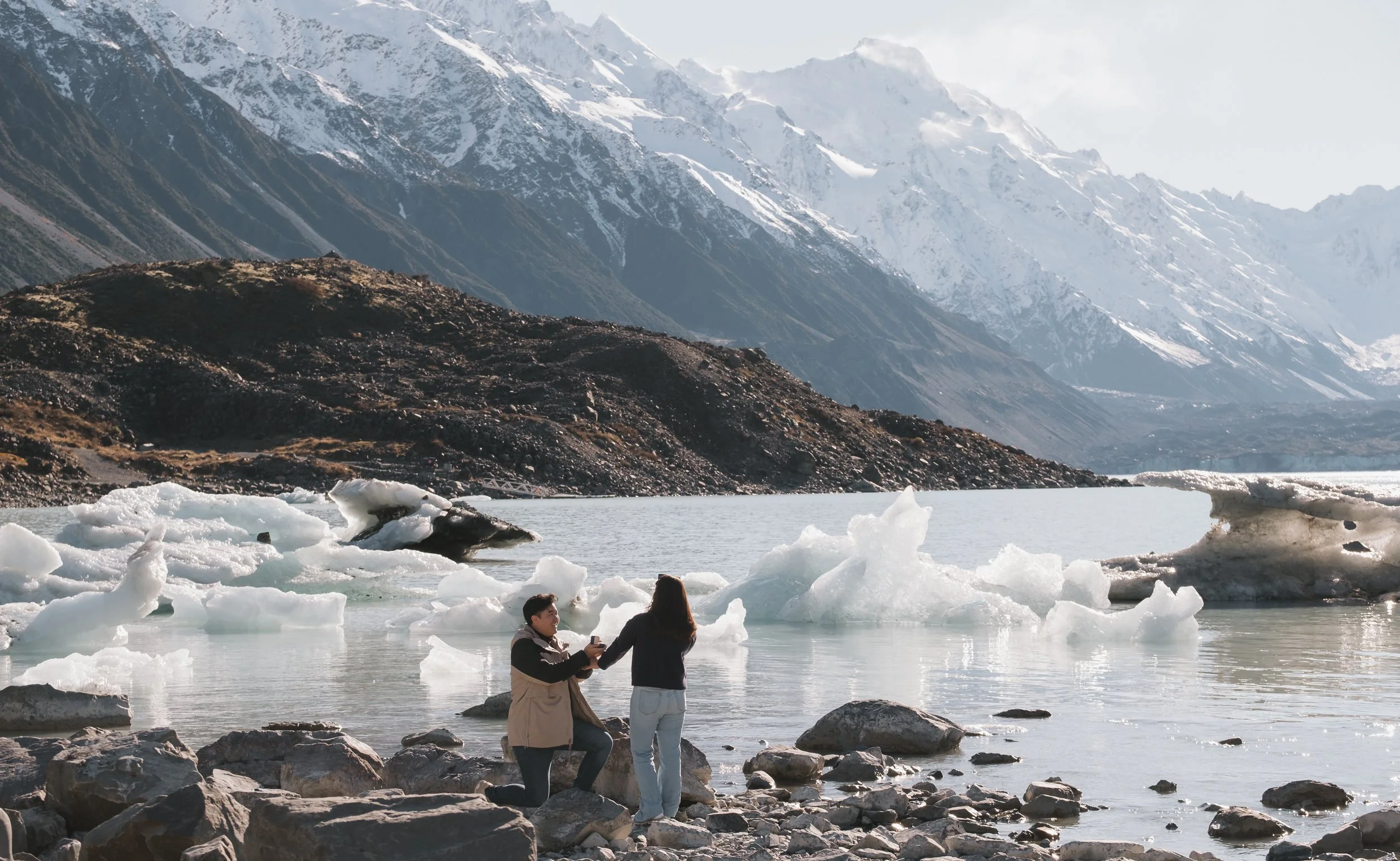 Two people stand near icy water with snow-covered mountains in the background.