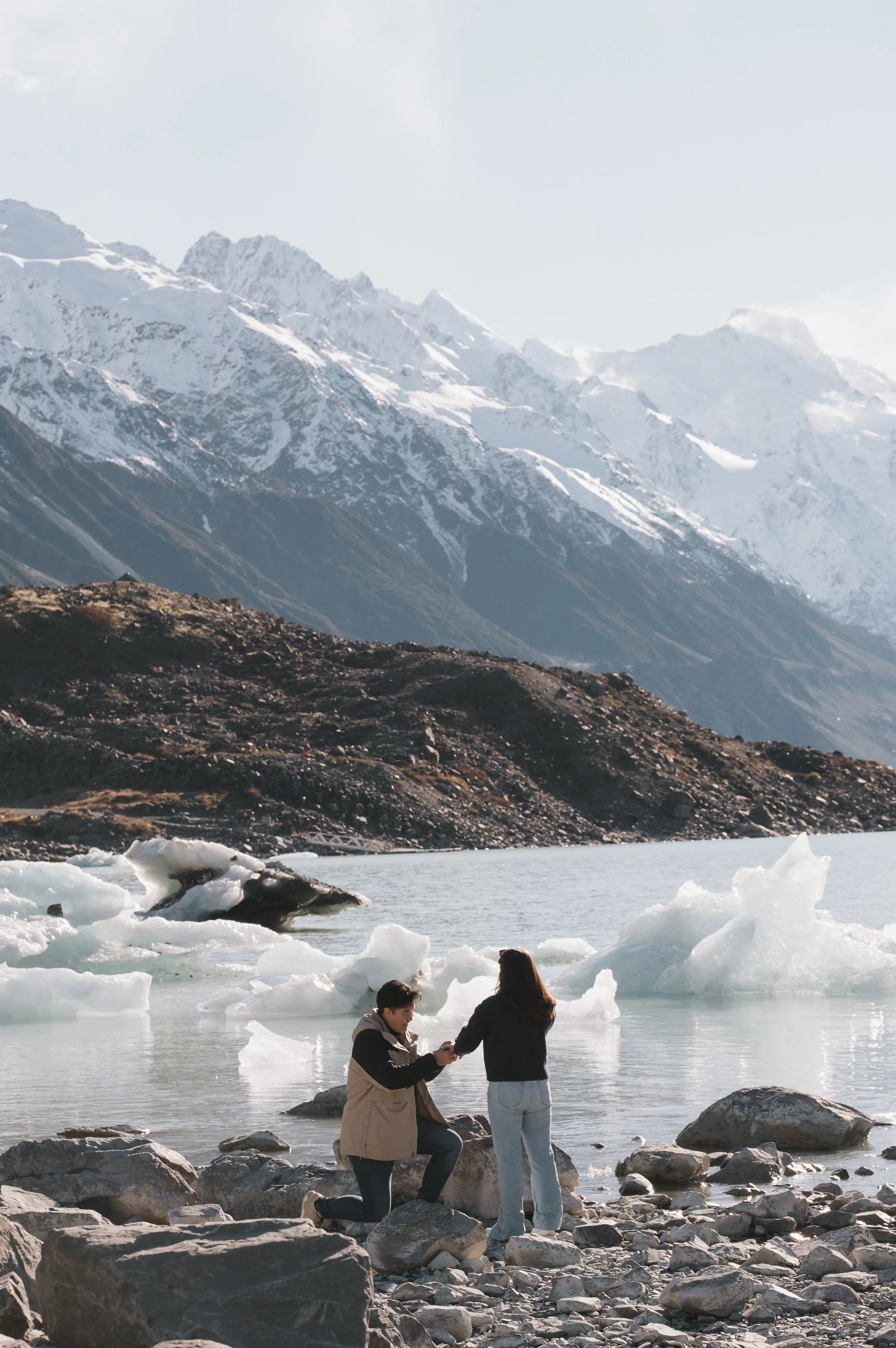 Two people are standing in icy water at a rocky lakeshore with snow-capped mountains in the background. One person is kneeling and proposing to the other, who has long hair and is wearing a white and blue jacket and light jeans. The person kneeling i