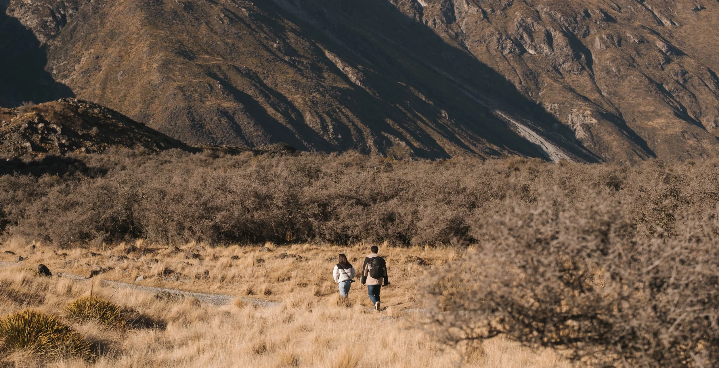 Two people walking through a field of dry grass towards a mountain range with barren, rocky slopes and sparse vegetation.