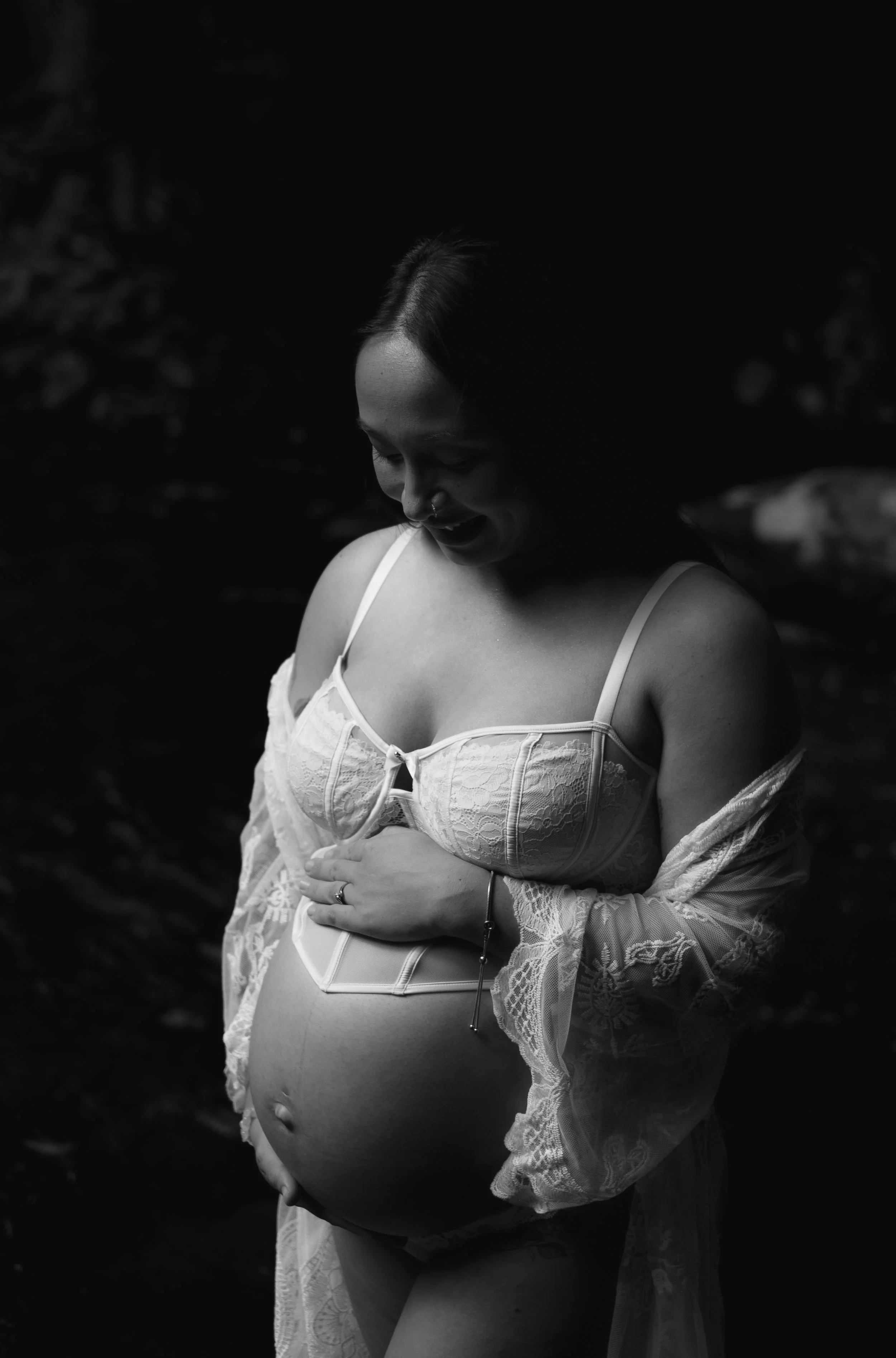A black and white photograph of a pregnant woman smiling with her head tilted downward, wearing a lace top and beside her hand resting on her belly.
