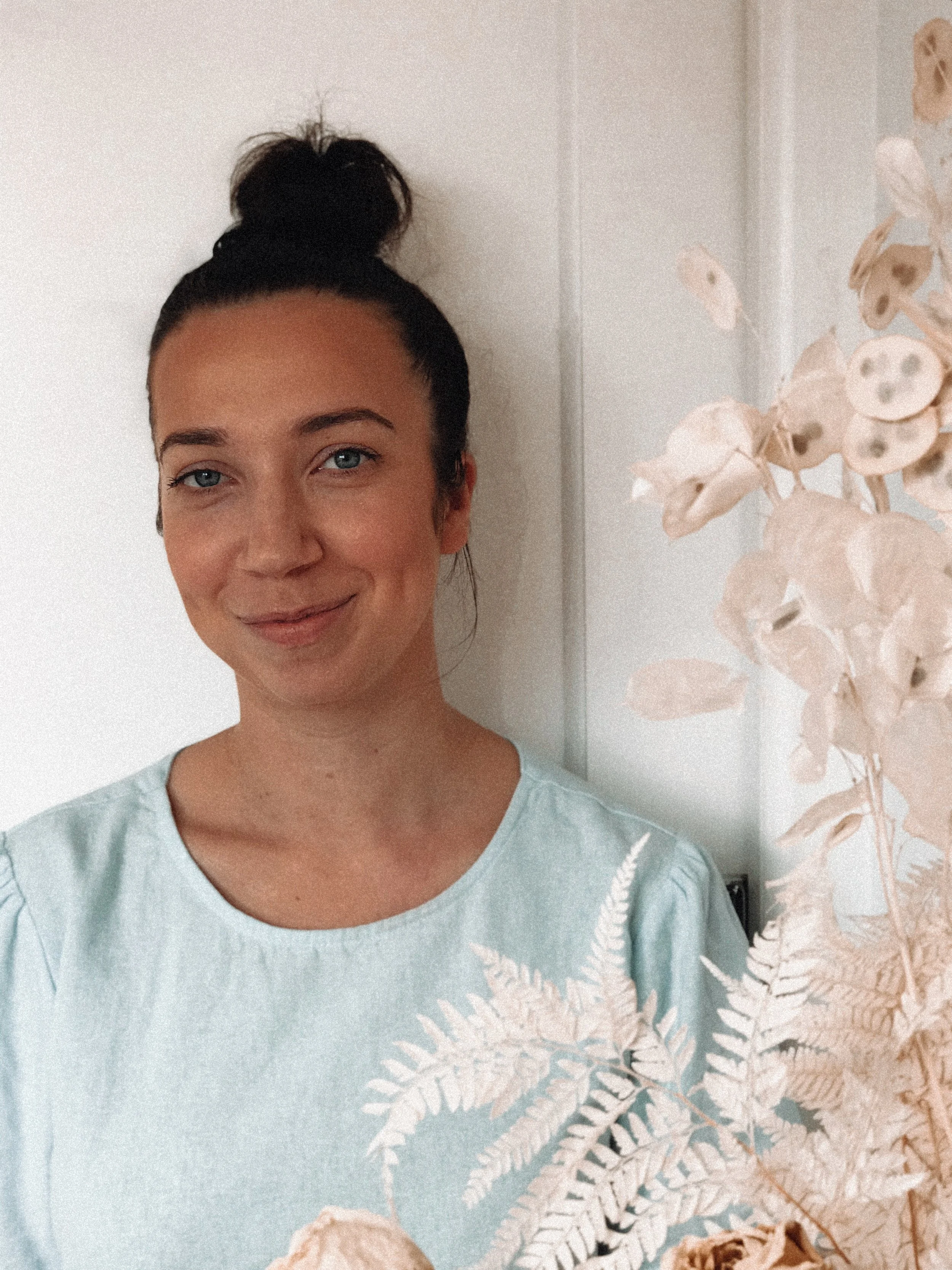 Woman with hair in a bun wearing a light blue shirt next to dried floral arrangement and white door background.