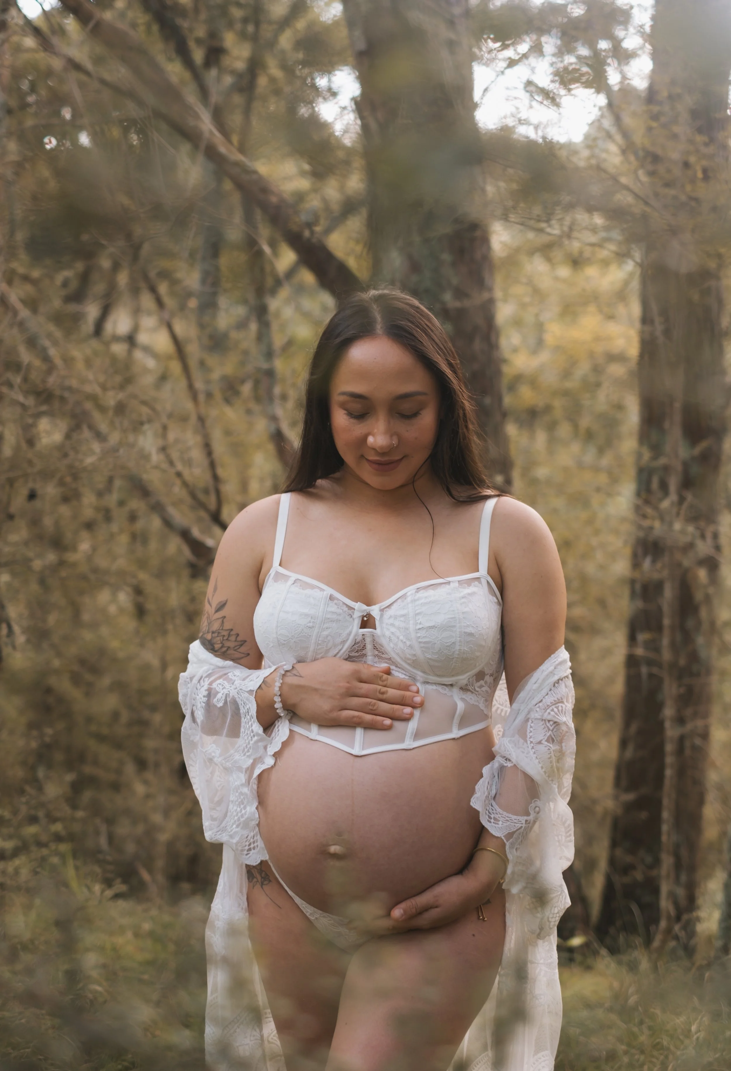 A pregnant woman standing in a forest wearing white lingerie and a lace robe, holding her pregnant belly with one hand and resting her other hand on her chest, looking down with a serene expression.