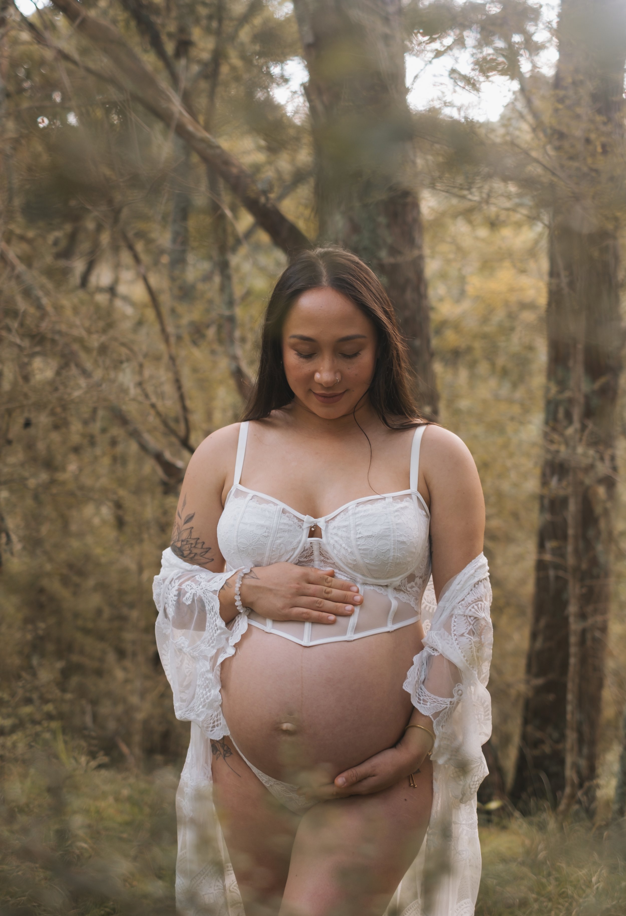 A pregnant woman standing in a forest wearing white lingerie and a lace robe, holding her pregnant belly with one hand and resting her other hand on her chest, looking down with a serene expression.