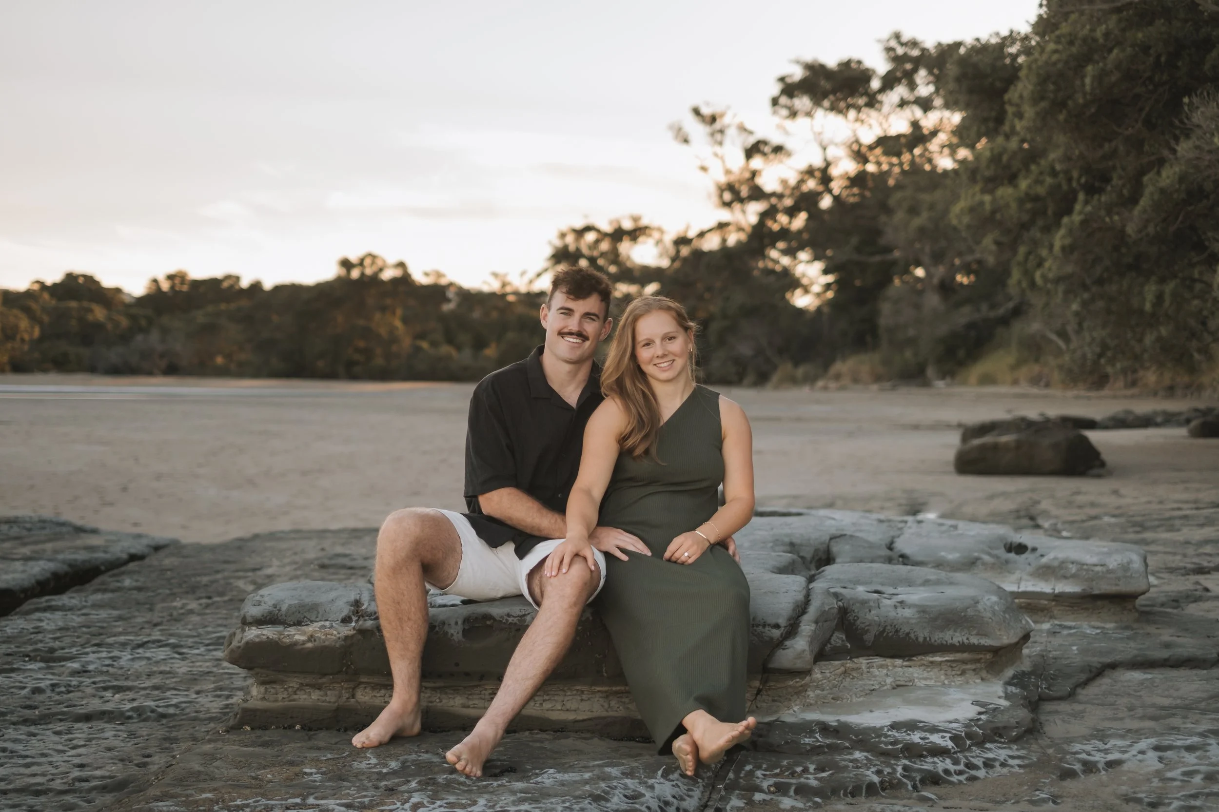 A man and woman sitting on rocks near the water's edge on a beach, smiling at the camera with trees in the background during sunset.