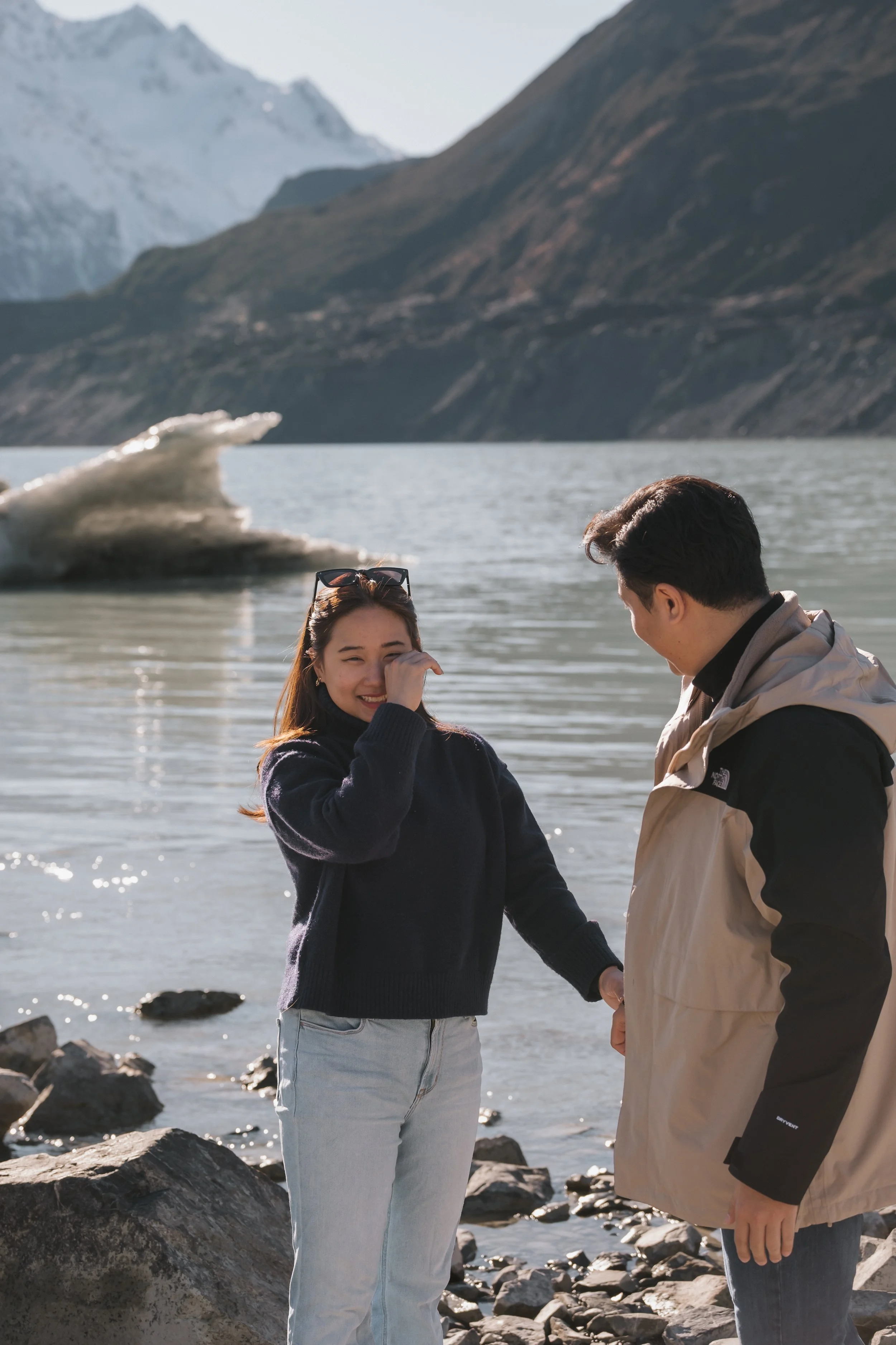 A man and woman standing by a lakeshore holding hands, with a mountainous landscape and a glacier in the background.