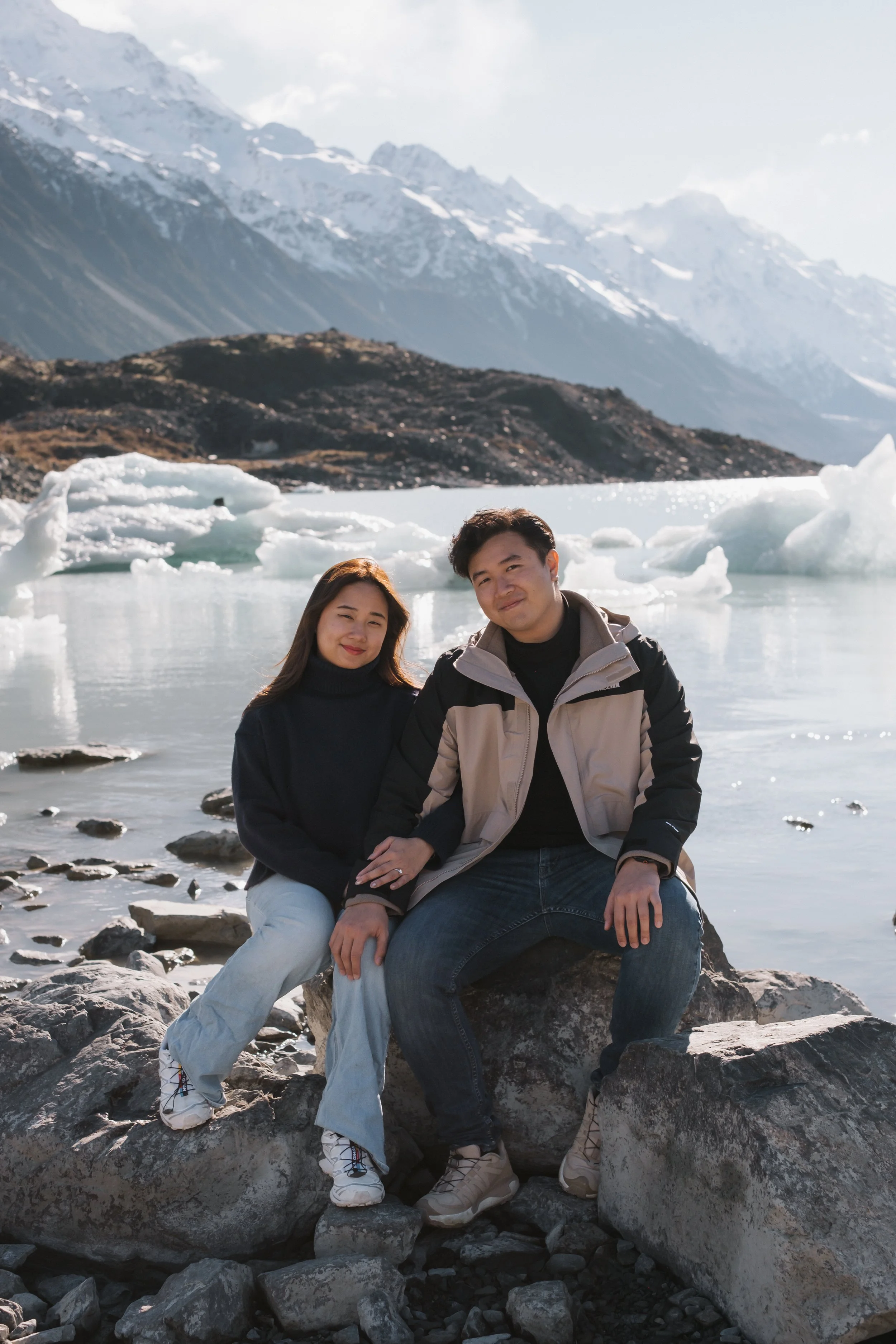 A couple sitting on rocks by a lake with icebergs, mountains with snow, and cloudy sky in the background.
