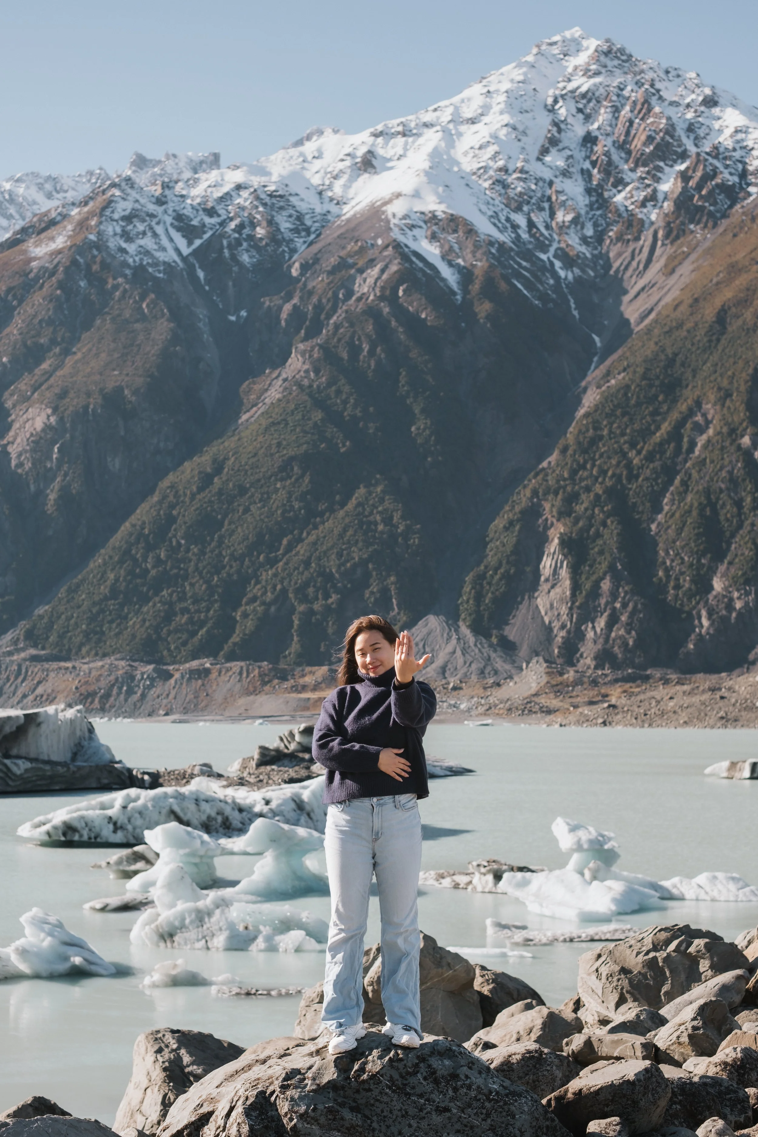 A woman standing on rocks near icy water with snow-capped mountains in the background, waving.