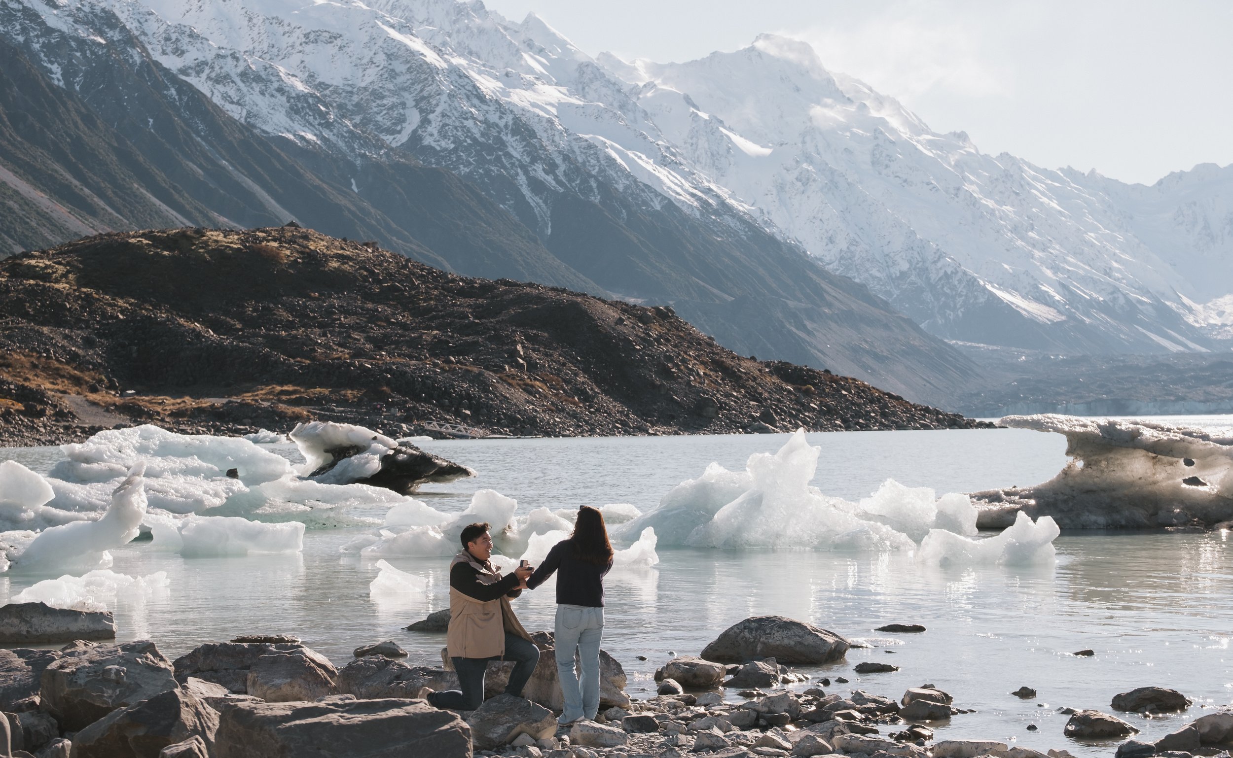 A couple standing in icy water holding hands with mountains and snow in the background.