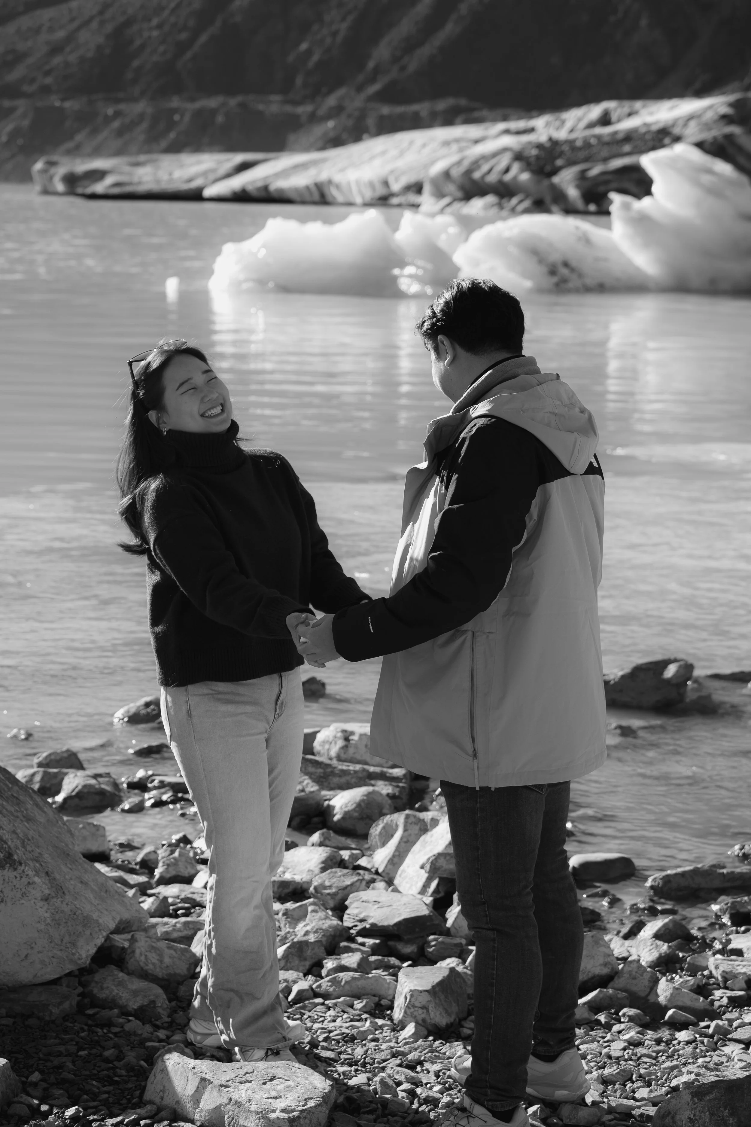 A man and woman holding hands and smiling at each other on a rocky lakeshore with icebergs and mountains in the background.