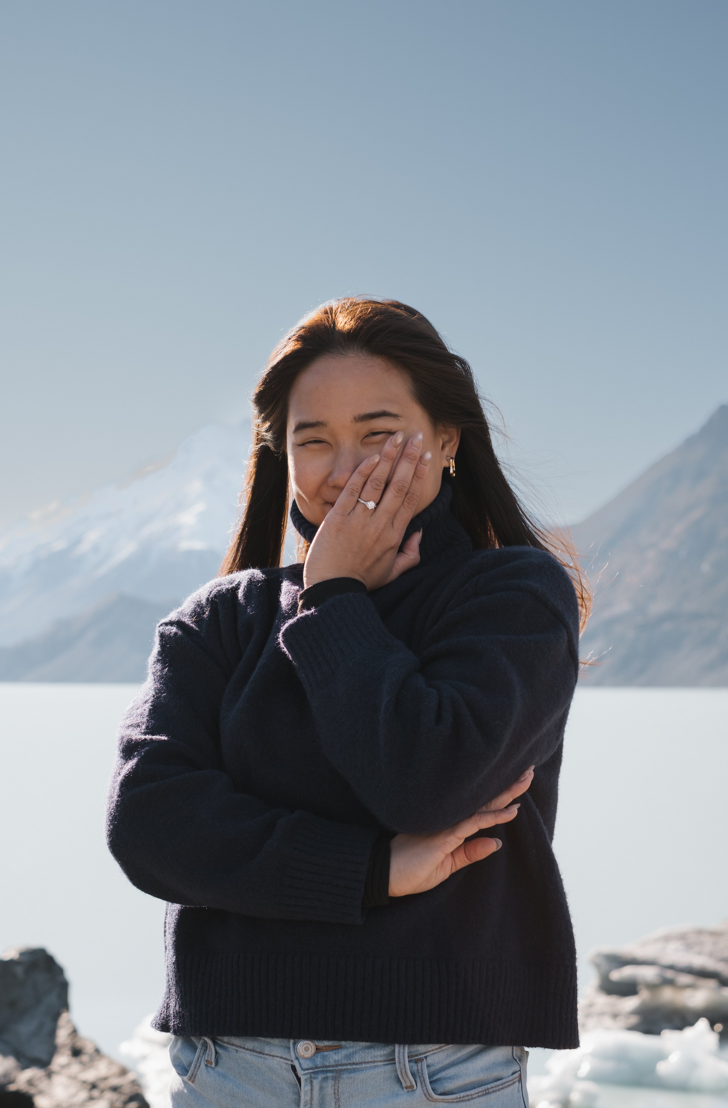 Young woman with long brown hair, wearing a dark sweater and light blue jeans, standing outdoors near snow-covered mountains, covering her face with her hand and smiling.