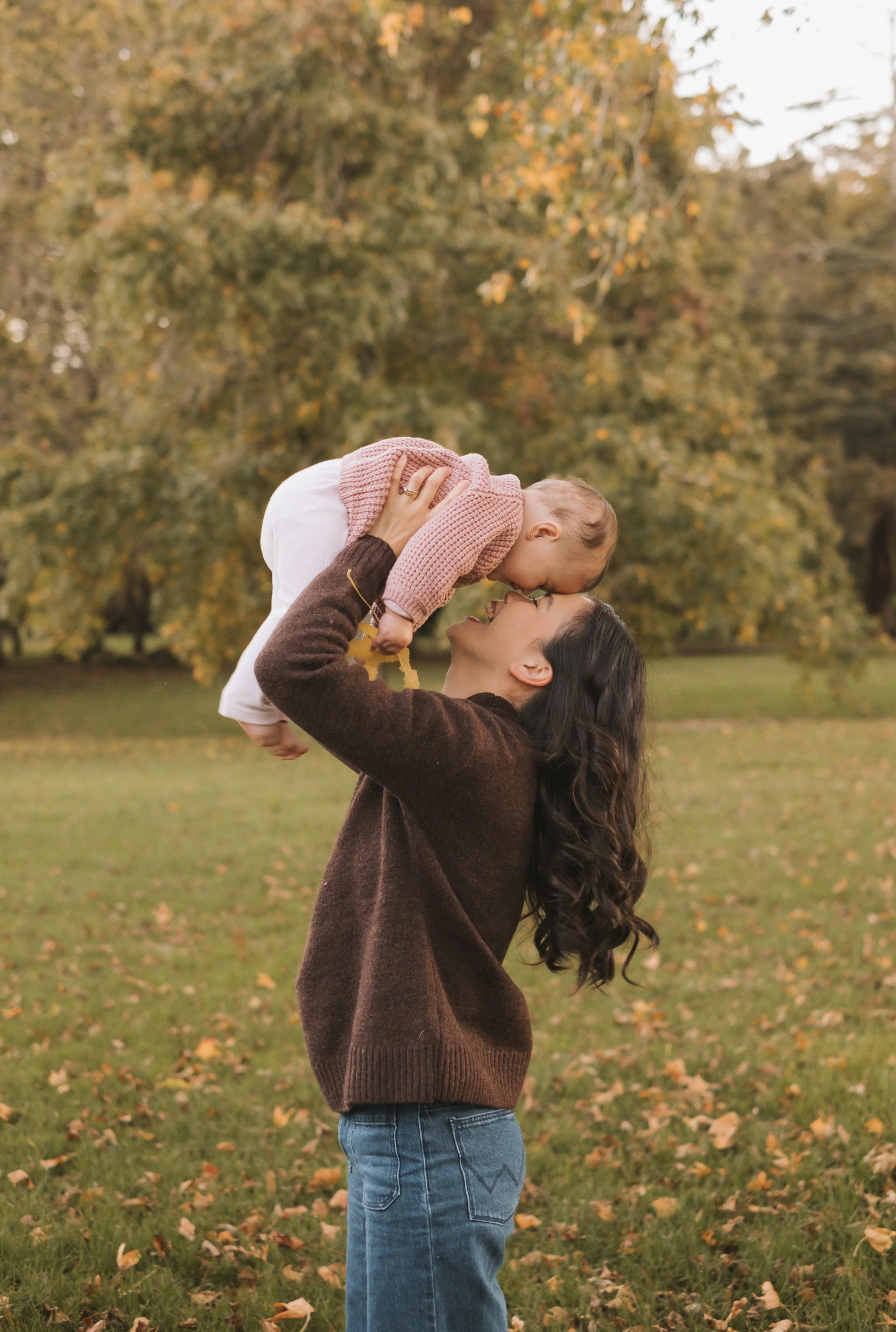 A woman with long dark hair and a brown sweater is holding a toddler in the air outdoors. The toddler is dressed in a pink sweater and white pants. The scene takes place in a park with green grass and fallen autumn leaves, and trees with yellow and o