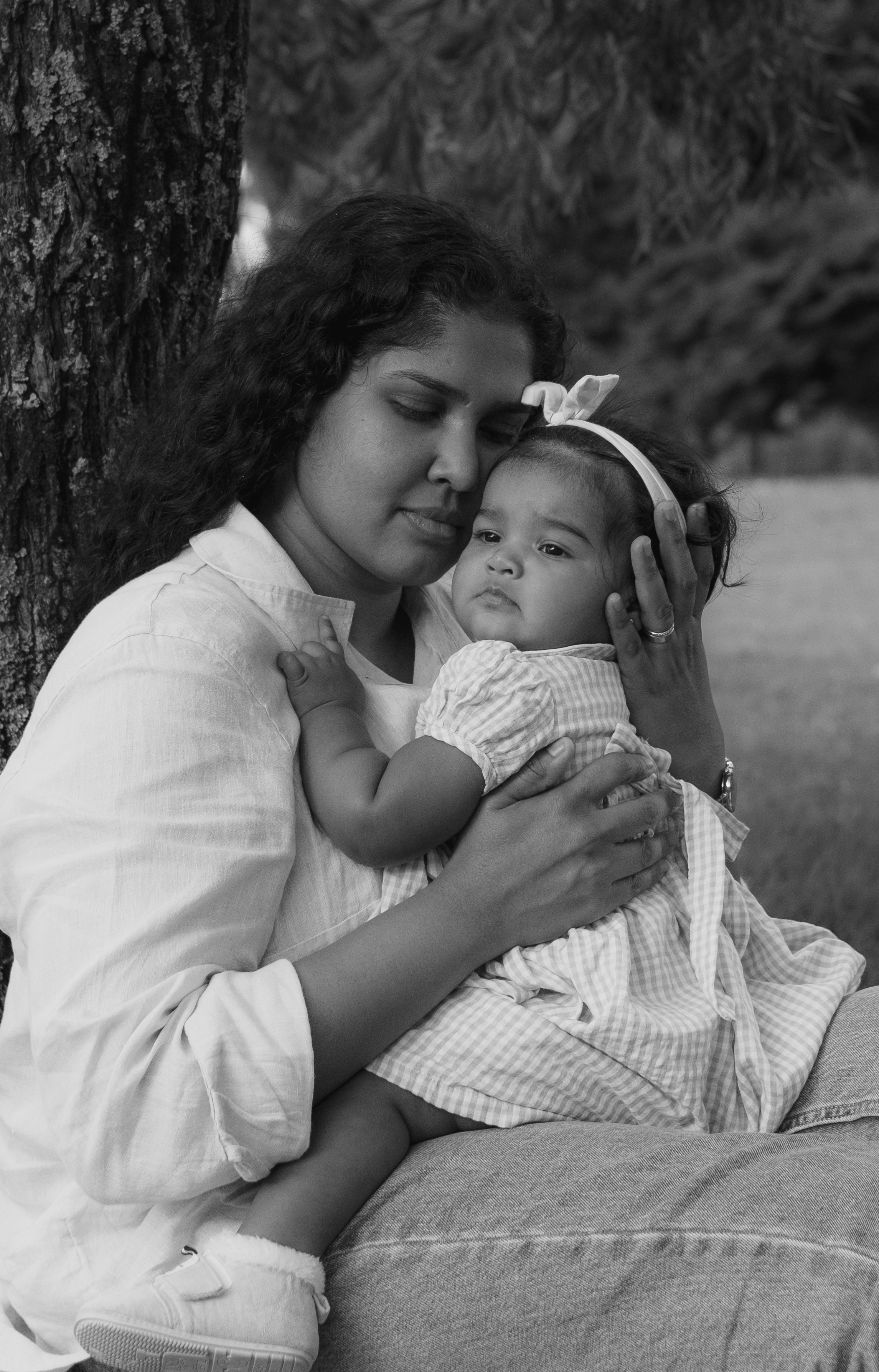 A woman with curly hair and a young girl with a headband are sitting outdoors near a tree. The woman is holding the girl close and gently resting her head against the girl's forehead with her eyes closed, creating a tender and loving moment. The girl