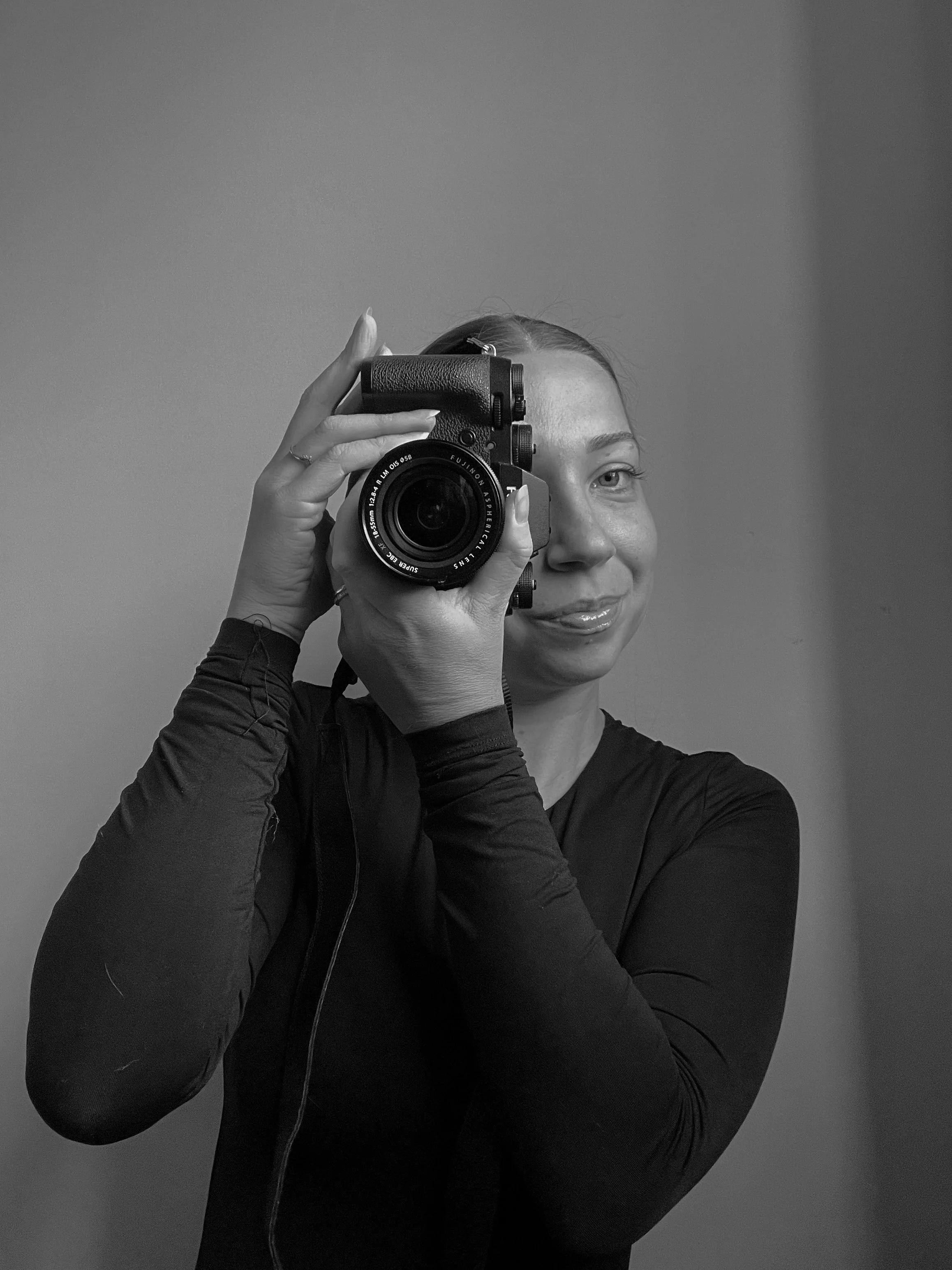 Black and white photo of a person taking a picture with a camera, wearing a long-sleeve top.
