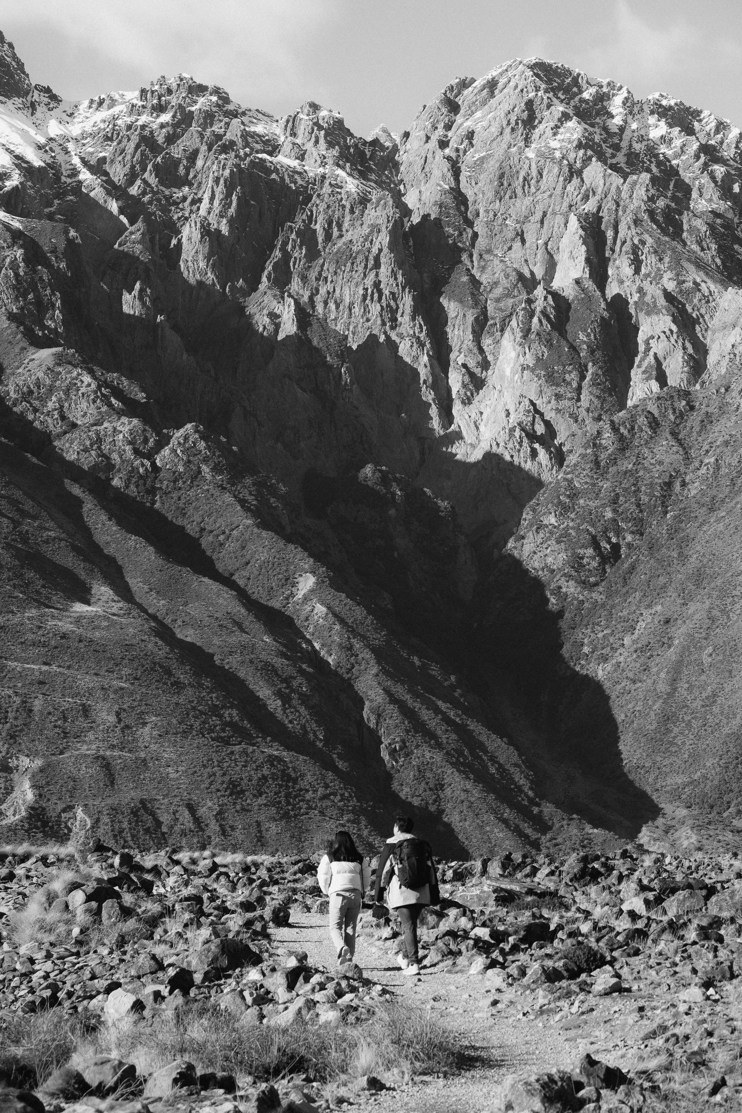 Two hikers walking on a rocky trail towards large mountains with steep, jagged peaks. The landscape is rugged and appears to be in a desert or arid environment, with sparse vegetation and clear sky.
