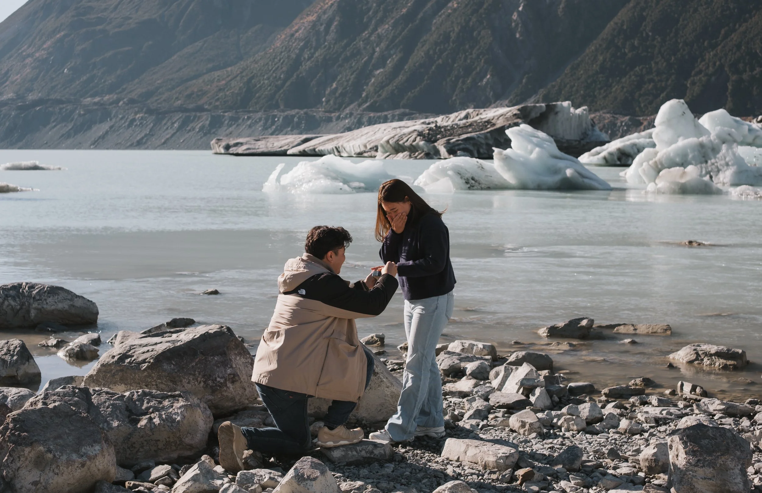 A man is proposing marriage to a woman on a rocky shoreline with ice chunks and glaciers in the background.