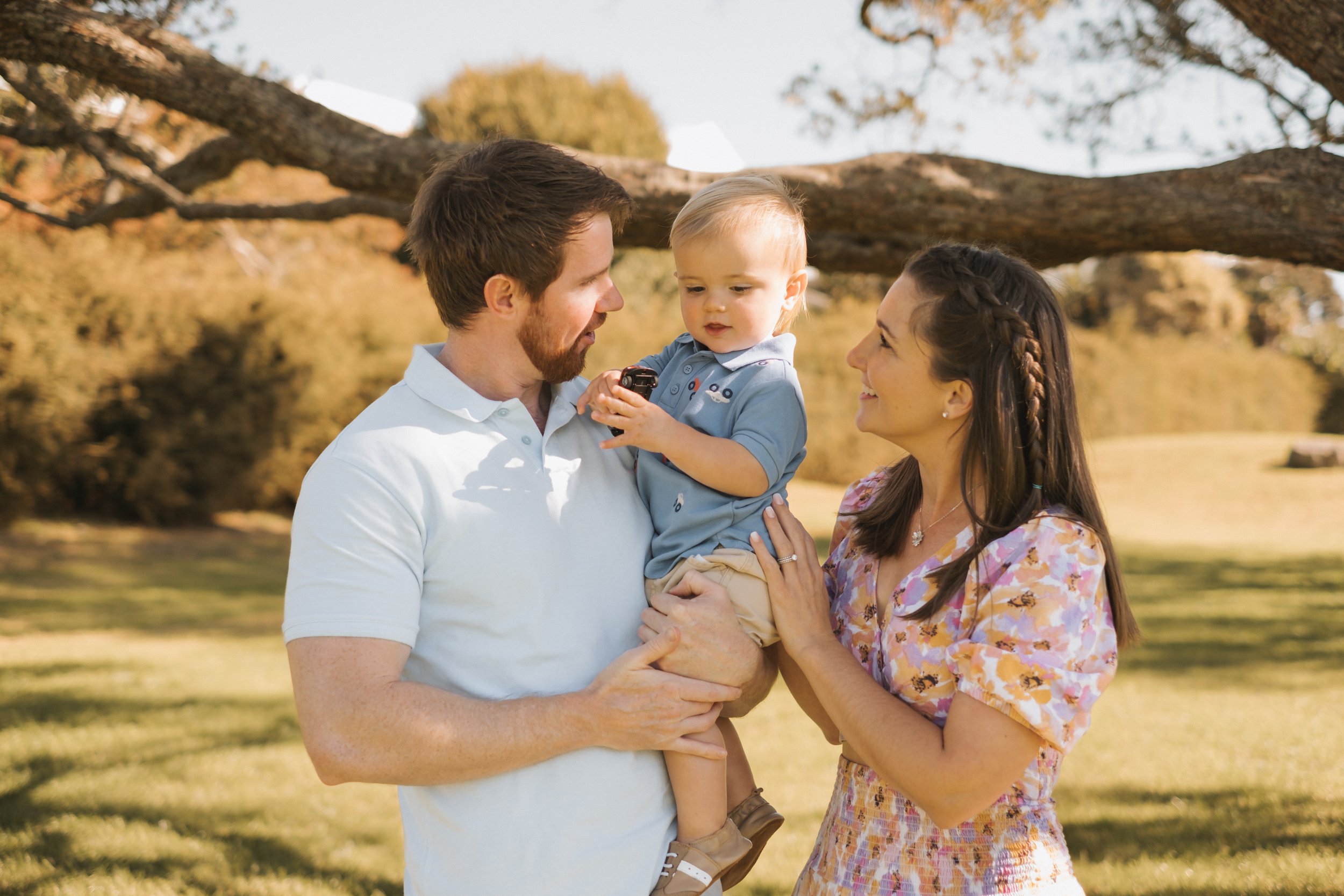 A family standing outside in a park on a sunny day. A man in a light blue shirt holds a small child dressed in a blue outfit. A woman in a floral dress stands beside them, smiling at the child.