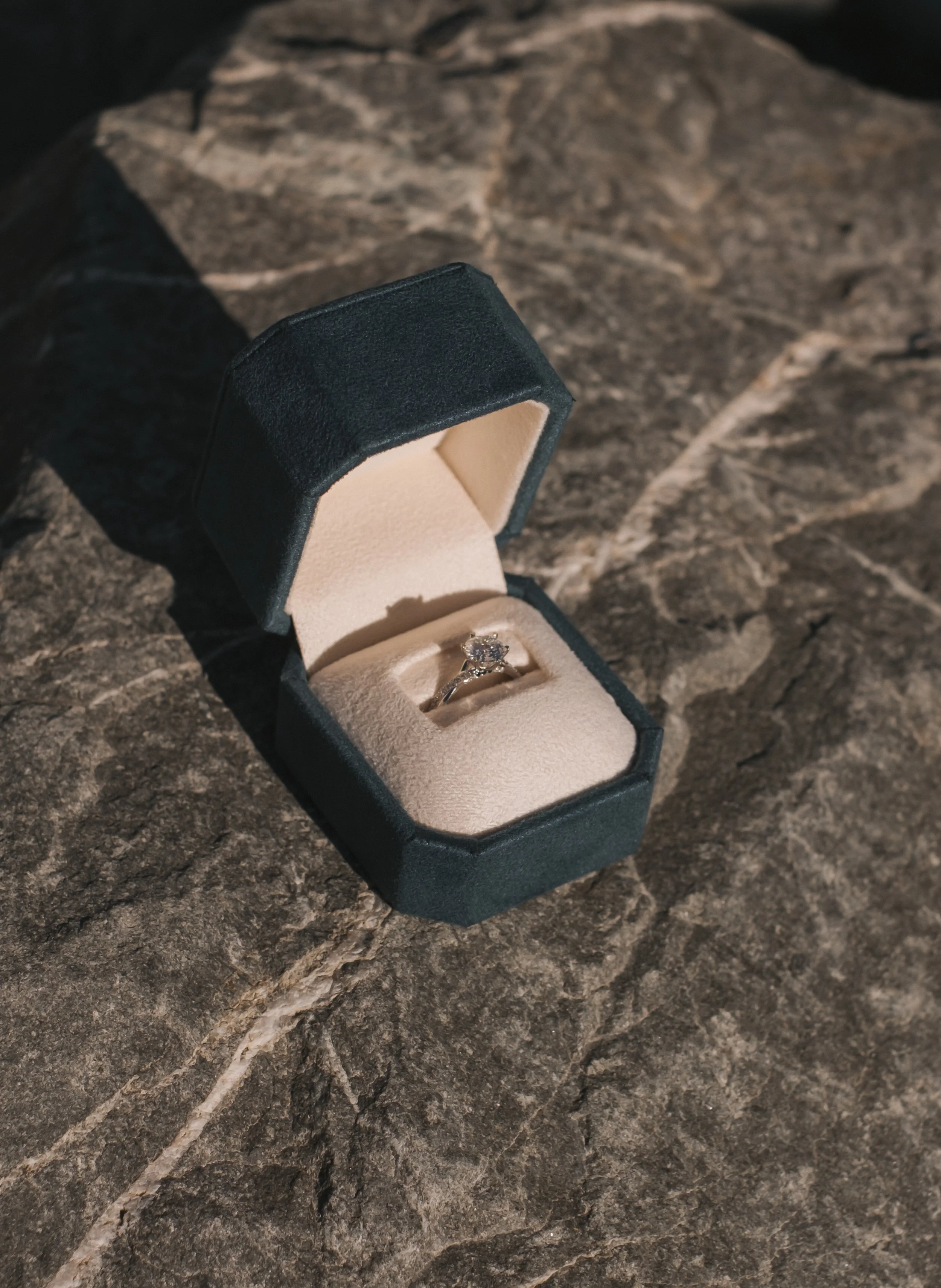 A silver engagement ring with a large diamond in a black jewelry box with a beige interior, placed on a dark textured stone surface.