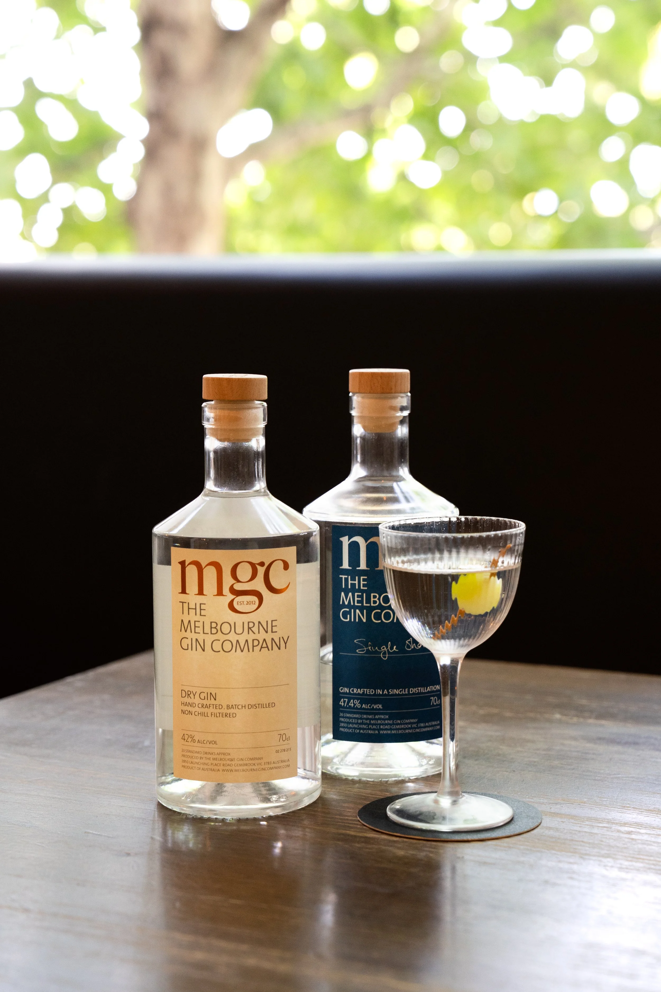 Two bottles of Melbourne Gin Company, one labeled Dry Gin and the other labeled Single Shot Gin, along with a filled cocktail glass on a wooden surface, with a blurred green leafy background.
