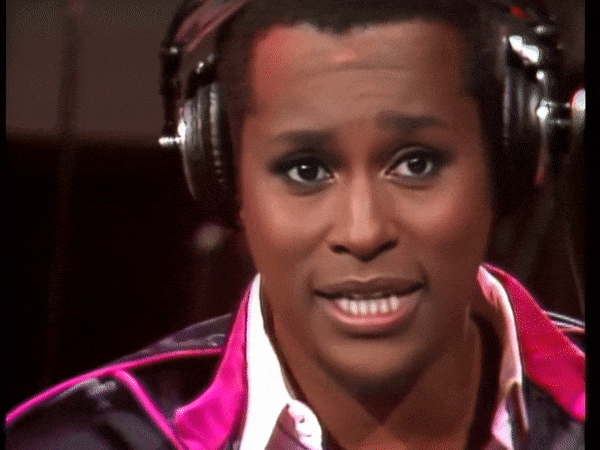 Person wearing headphones speaking into a microphone, dressed in a colorful shirt, animated expression.