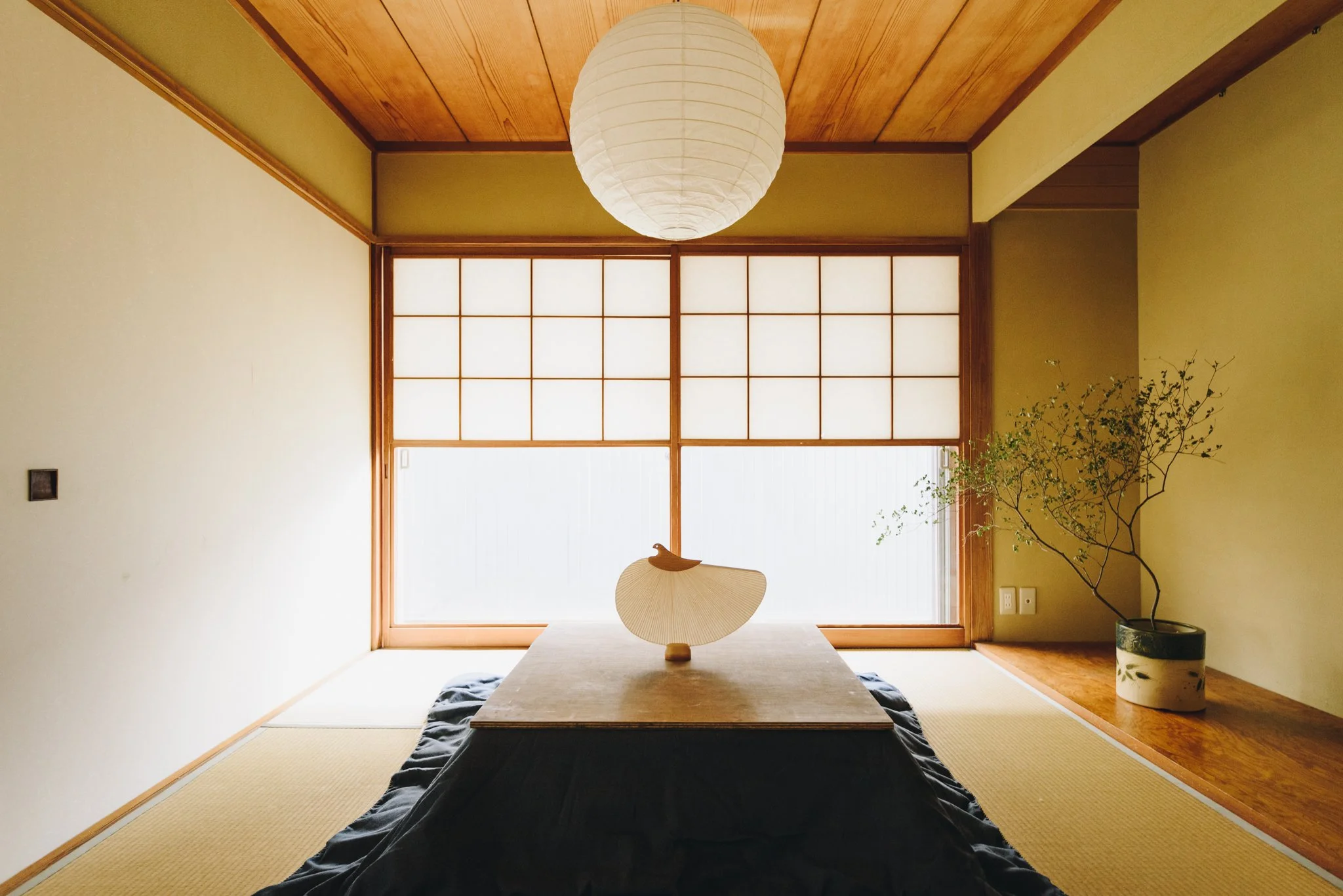 Minimalist Japanese-style room with tatami flooring, shoji sliding doors, a low wooden table with a decorative fan centerpiece, a hanging paper lantern, and a potted plant in the corner.