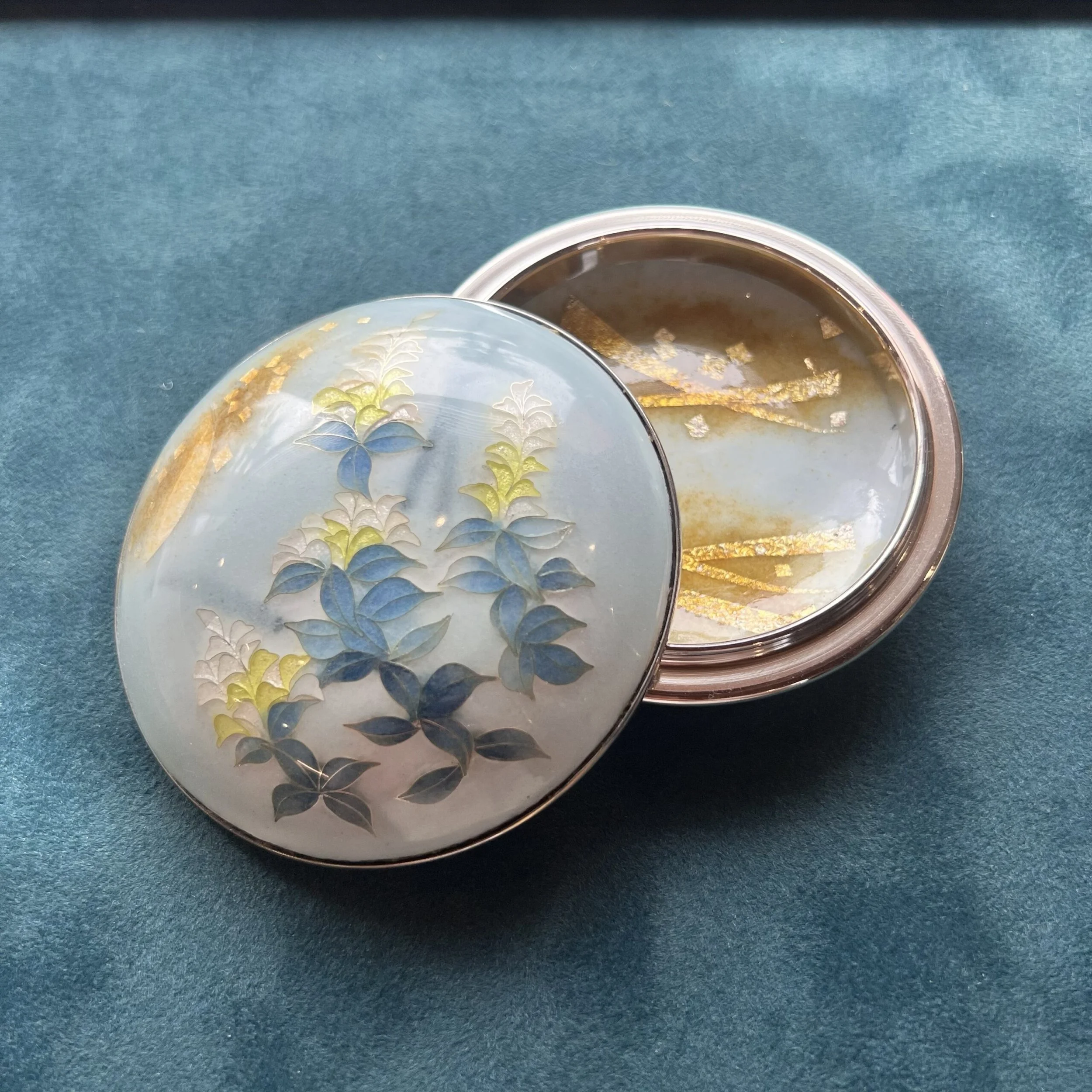 A decorative round container with a lid featuring a painted floral design, partially opened to reveal a jelly-like substance inside, resting on a blue fabric surface.
