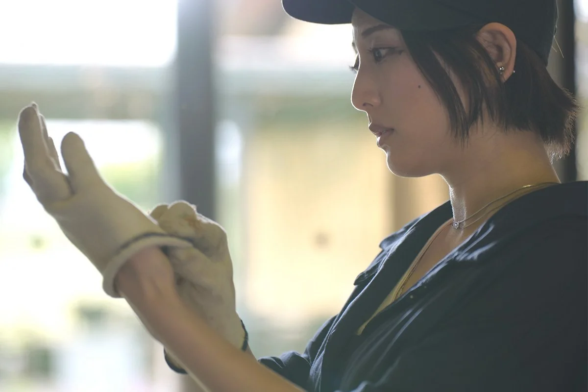 Kodawari. Tamura Shippo. A woman wearing a black cap and gloves is looking at her hand, adjusting her glove, inside a bright space with windows in the background.