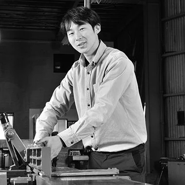 A man working at a table in a workshop or laboratory setting, smiling at the camera.