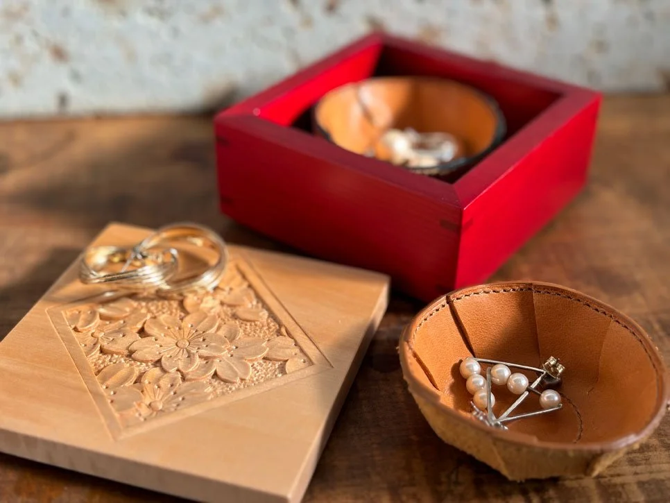 Kodawari. Oshima Kobo. A wooden box with a floral carving on top, a red box with a black interior containing jewelry, and a small leather bowl holding pearl and diamond jewelry on a wooden surface.