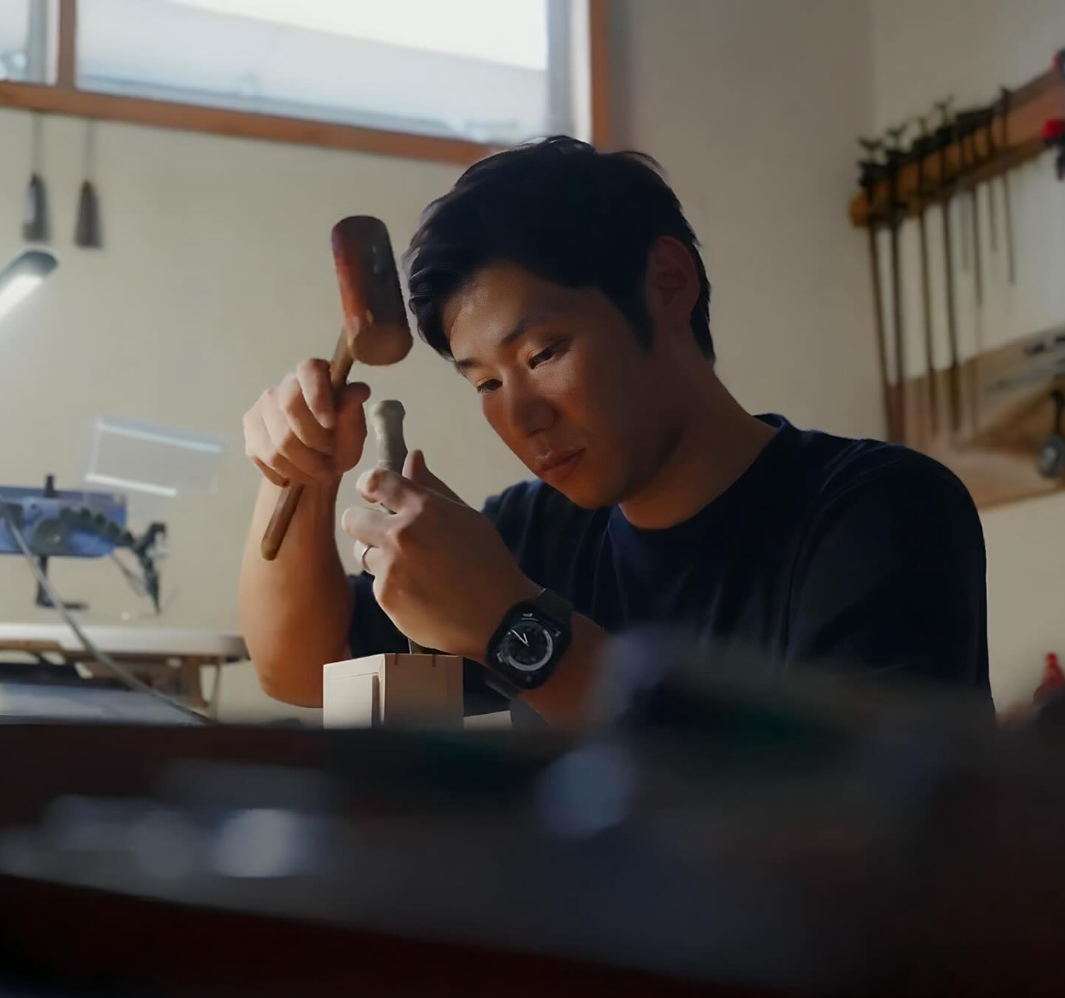 Kodawari. Eisui Oshima. A man with dark hair, wearing a black shirt and a watch, is working on a wooden piece with a chisel and hammer in a workshop. There are tools and equipment in the background.