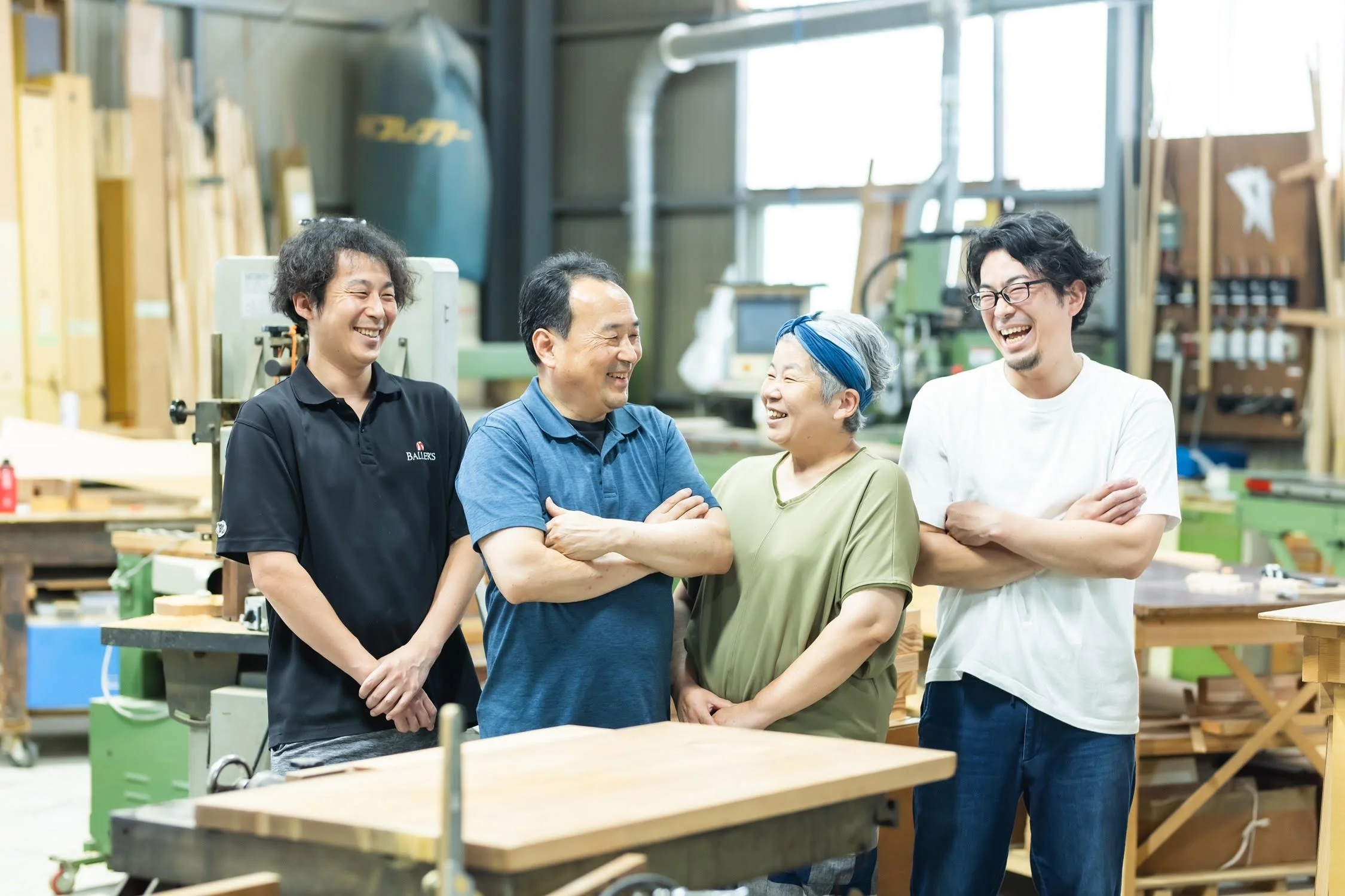 Four people laughing and talking in a woodworking workshop. Tools and wood materials are visible in the background.