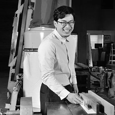 A man with glasses smiling and working with wood in a workshop.