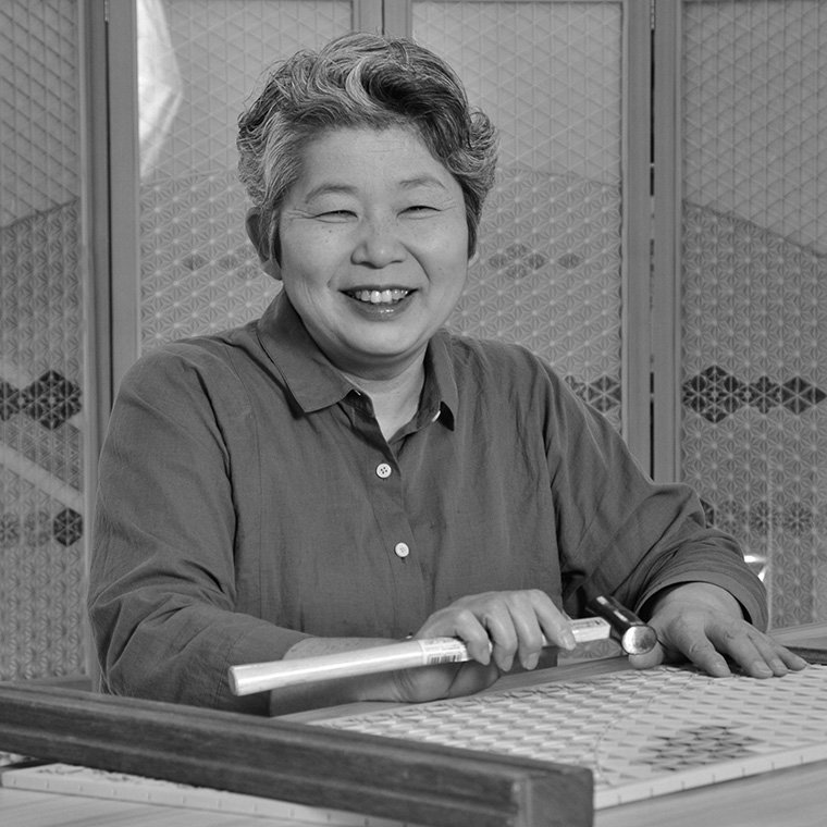 A smiling woman with short curly hair playing shuffleboard, holding a cue stick, in an indoor recreational area.