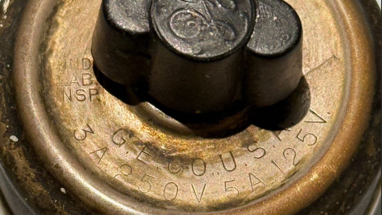 Macro close-up of a 1910s GE rotary snap switch flat dial, showing the classic cursive General Electric logo and original voltage ratings stamped into the aged brass dome.