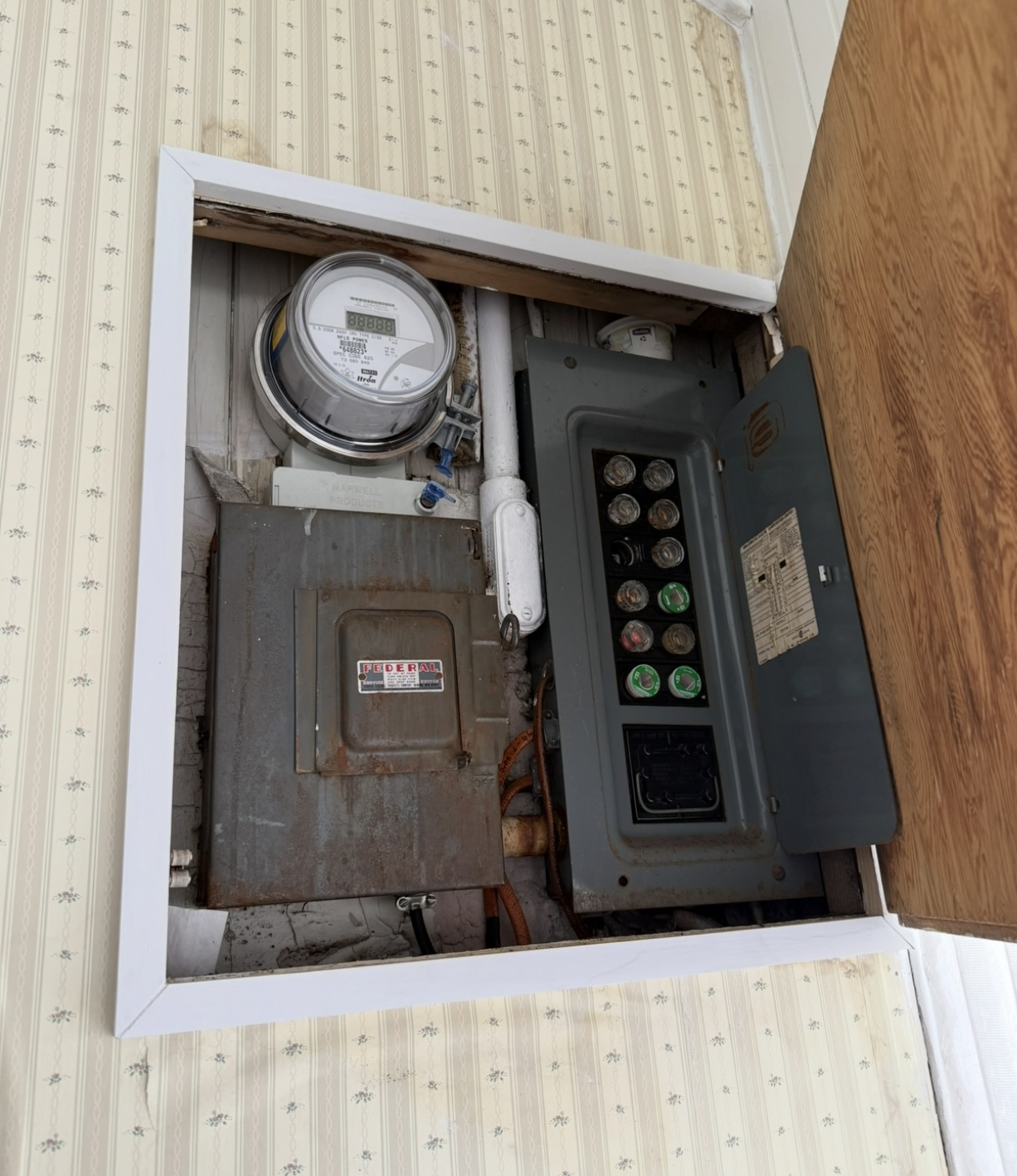 A mid-century indoor electrical setup hidden inside a wooden wall cabinet in a historic St. John's home, featuring a modern utility meter mounted above a vintage Federal disconnect switch and an obsolete panel utilizing Edison-base glass plug fuses.