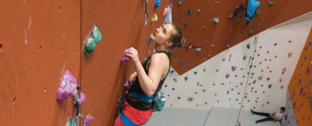 Kvinne klatrer på en innendørs klatrevegg med fargerike klatregrep. Personen er sikret med tau, og det er flere klatrere i bakgrunnen på en lignende vegg.