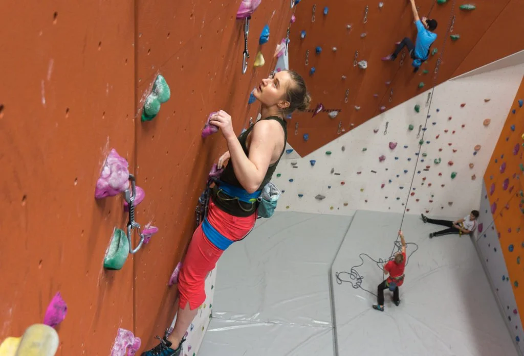 Kvinne som klatrer på en innendørs klatrevegg med fargerike håndtak, med andre klatrere i bakgrunnen.
