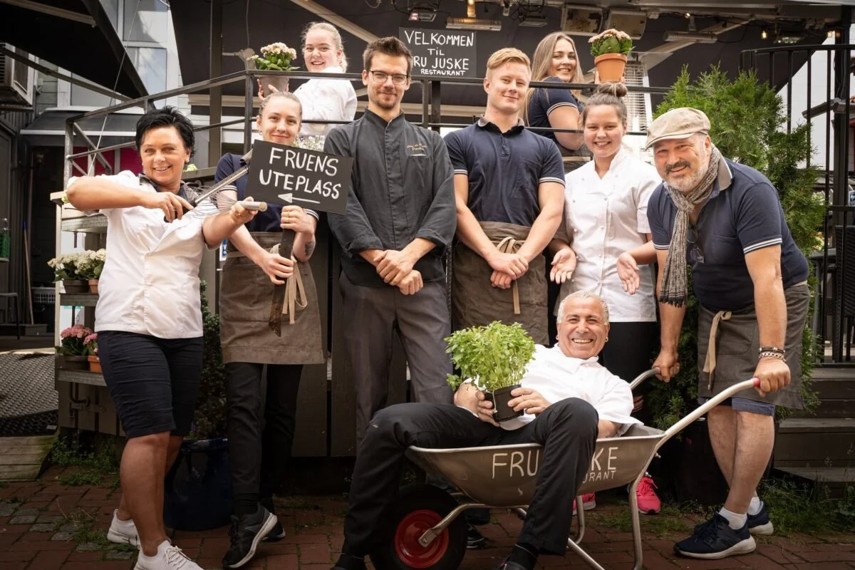 Gruppebilde av restaurantansatte, med en kvinne som holder et skilt som sier "Fruens uteplass". Flere personer smiler, inkludert en mann som sitter i en trillebår og holder en plante. Bakgrunnen viser velkomstskilt for "Fru Juske".