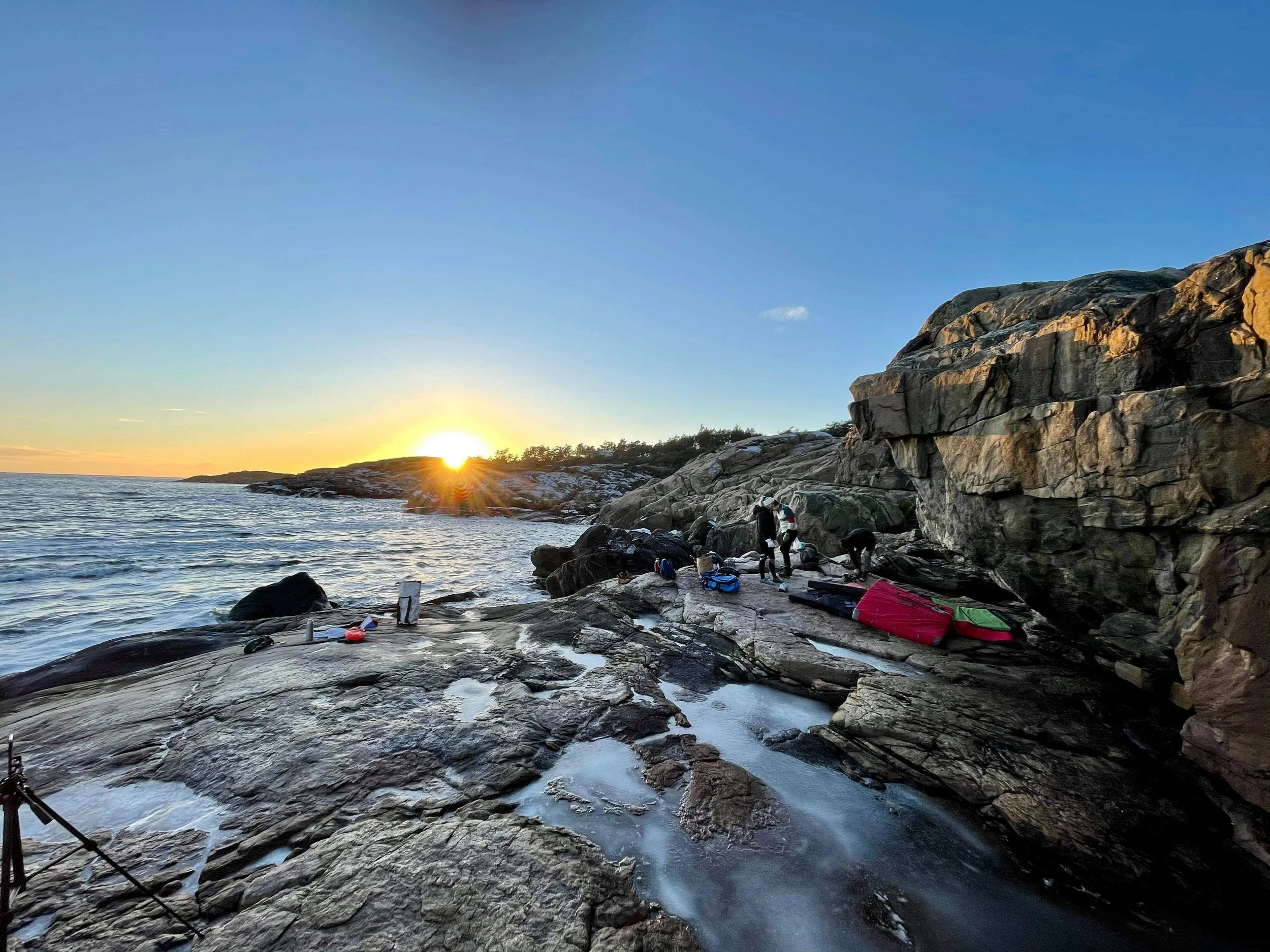 Solnedgang ved kysten med personer og kajakker på en steinete strand.