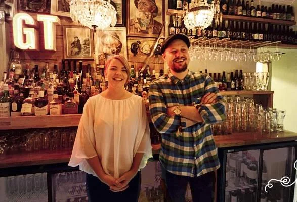 To personer står bak bardisken. Det er mange flasker med alkohol og glass på hyller bak dem. Det er også dekorasjoner som bilder og bokstavskiltet "GT". En person har på seg en rutet skjorte og caps, mens den andre har på lys topp.