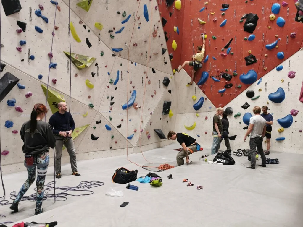 Personer klatrer på innendørs klatrevegg, med flere som forbereder seg eller hjelper til. Veggen har ulike fargede klatretak.