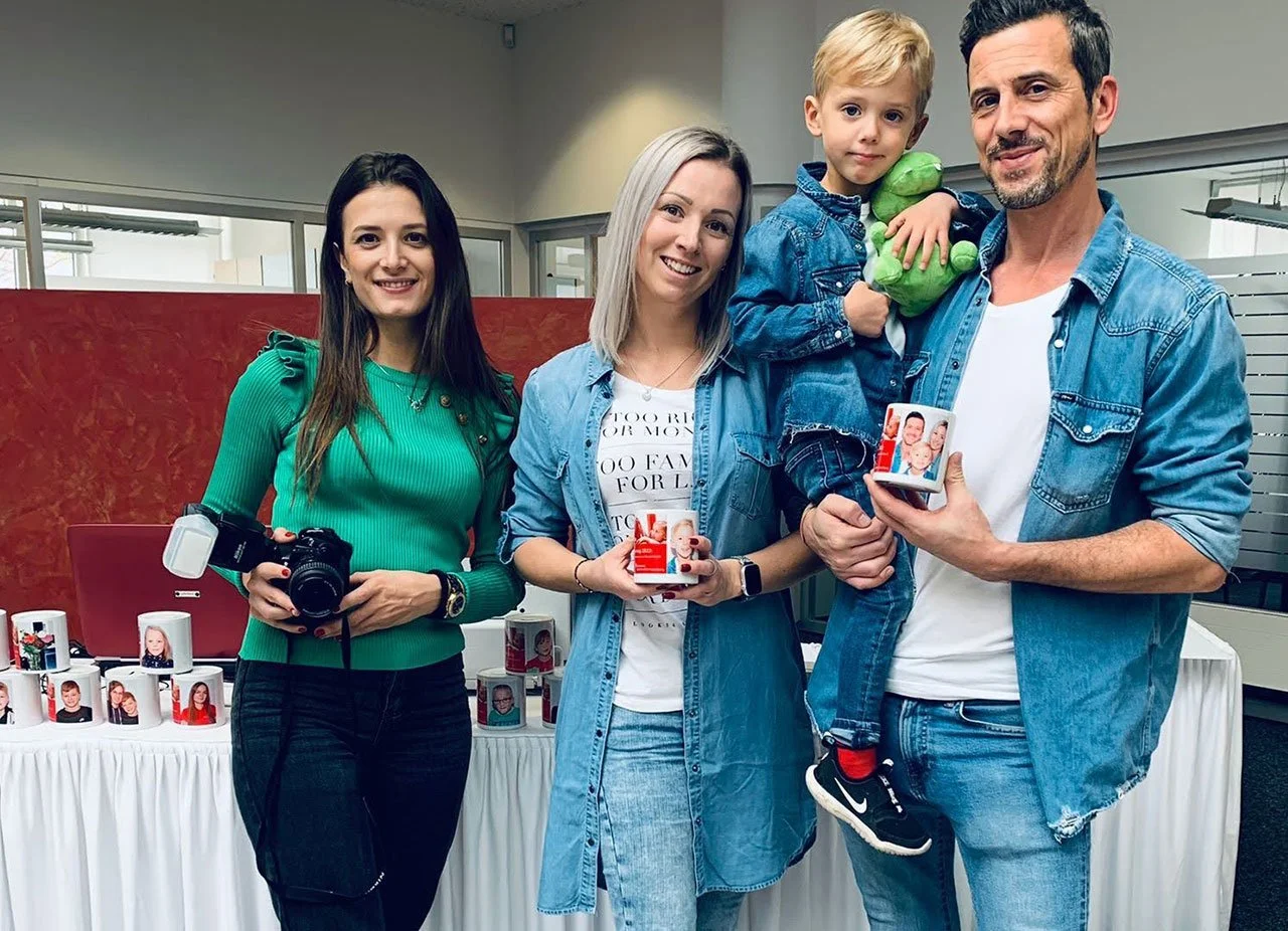Eine Gruppe von vier Menschen, drei Erwachsene und ein Kind, posieren in einem Raum. Das Kind hält einen grünen Teddybär, während die Erwachsenen kleine Fotogeschenke in den Händen halten. Im Hintergrund steht eine Tafel mit mehreren Fotomugs auf einem weißen Tisch.