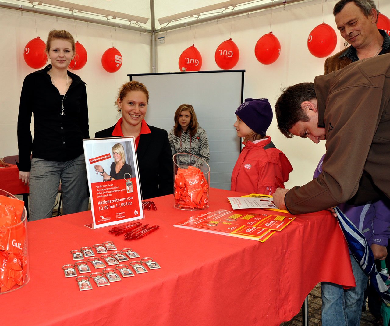 Menschen an einem Informationsstand mit roten Kugeln und einem Spendenkorb, es scheint eine Veranstaltung im Zusammenhang mit der Sparkasse zu sein, mit Personen, die Broschüren und Schlüsselanhänger erhalten.