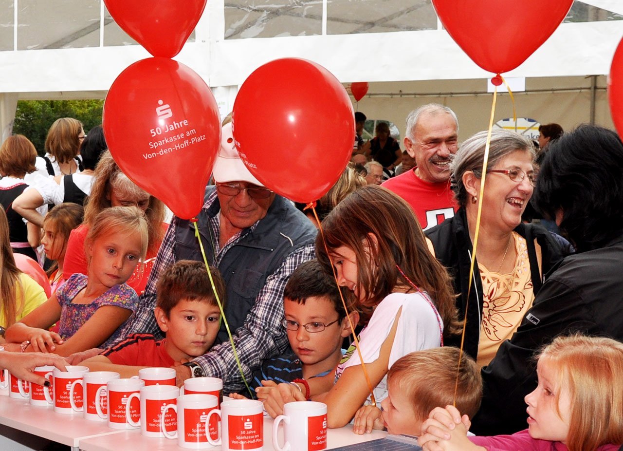 Viele Menschen feiern bei einem Fest unter einem weißen Zelt. Es gibt rote Ballons und Tassen mit dem Logo der Sparkasse. Einige Erwachsene und Kinder sind entspannt und lachen, einige schauen auf die Tische.
