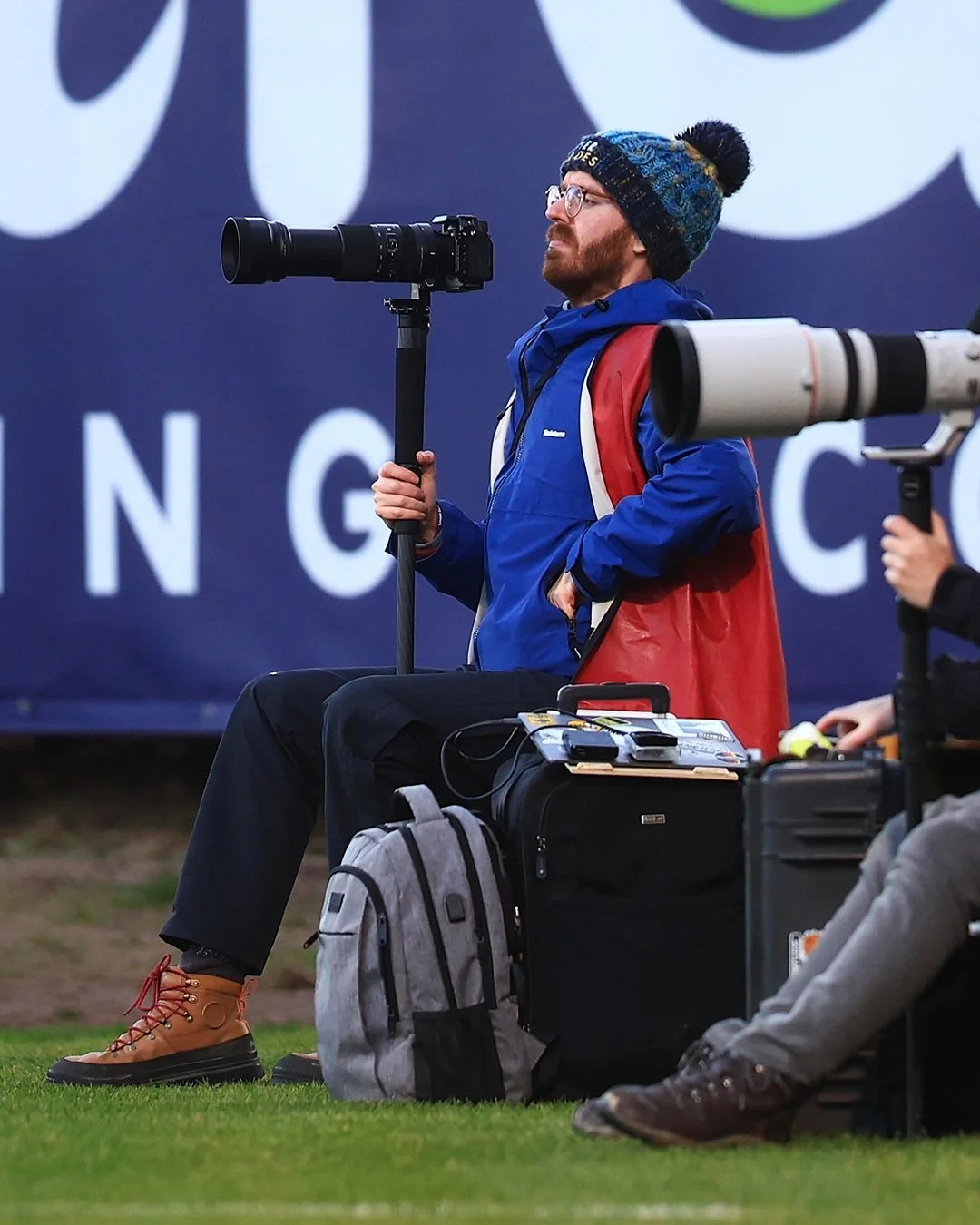Photographer seated with camera equipment, wearing a blue jacket and knit hat, holding a camera on a monopod.