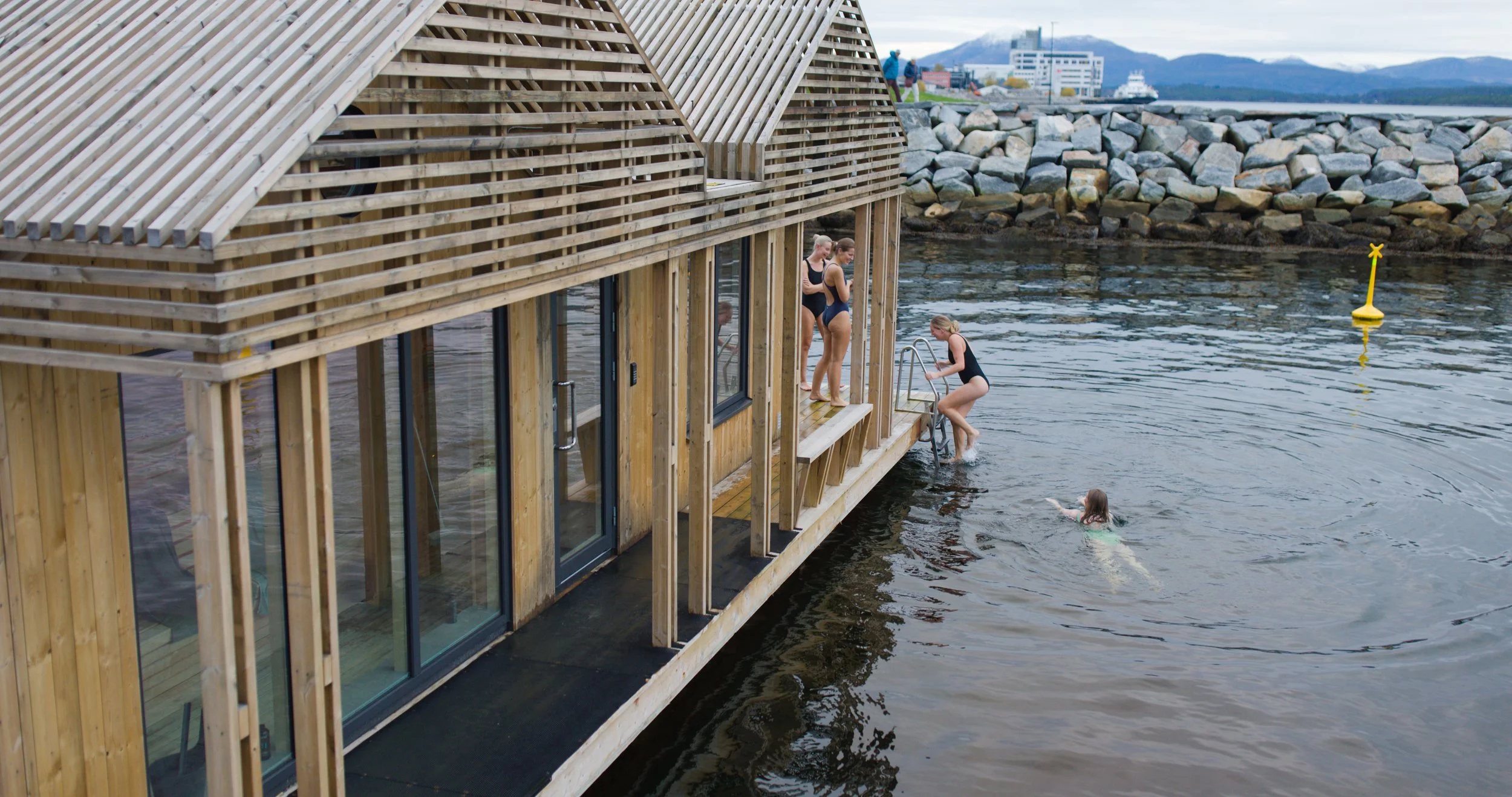 Fiskebrygge med flere barn som svømmer og deler en trehusliknende roterende badstue ved vannet, med fjell og bylandskap i bakgrunnen.